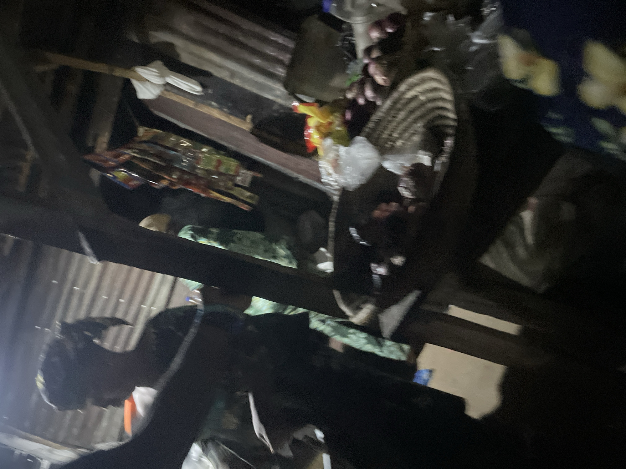 The image depicts a dimly lit scene, likely an outdoor market or roadside stall in Lokoja, Nigeria. In the foreground, a woven basket is filled with what appear to be red onions and other produce. To the left, a person's arm and shoulder are visible, partially obscuring the view. Behind the basket, a woman wearing a headscarf is seen in profile, with her body turned away from the camera. She is wearing a dark-colored garment. In the background, there are shelves displaying various items, including packets of snacks or food products. The overall environment suggests a humble commercial setting.