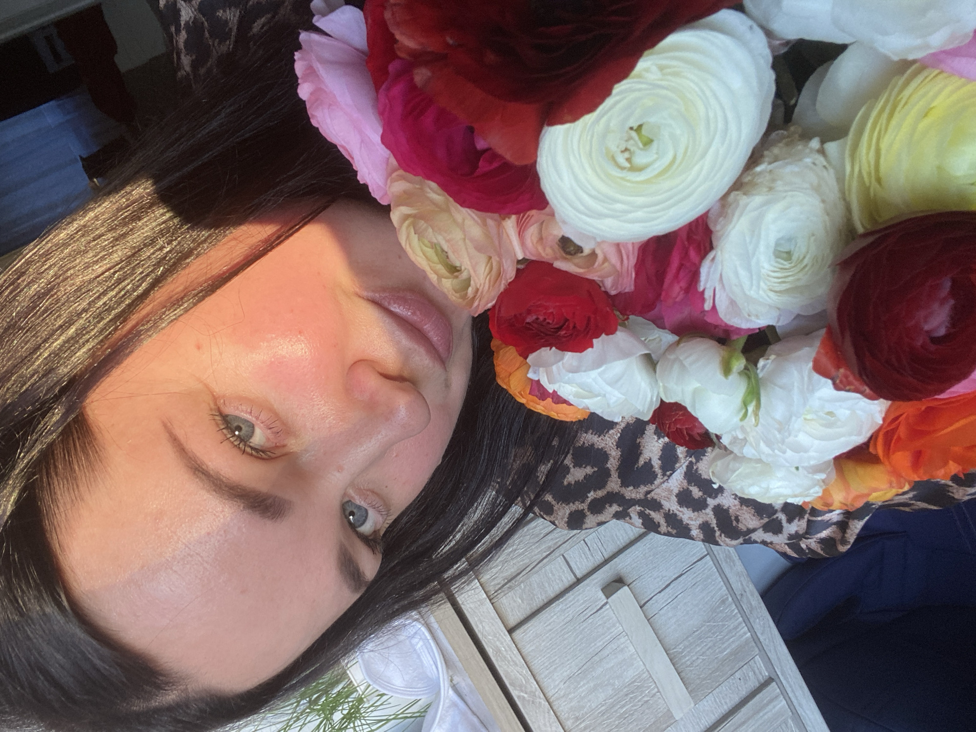 The image shows a woman's face surrounded by a bouquet of ranunculus flowers in various colors, including red, white, pink, yellow, and orange. The woman has dark hair and light-colored eyes. She is wearing a leopard-print top. The background includes a light-colored wooden surface and a glimpse of green foliage. There is no indication of the location being Sochi, Russia.