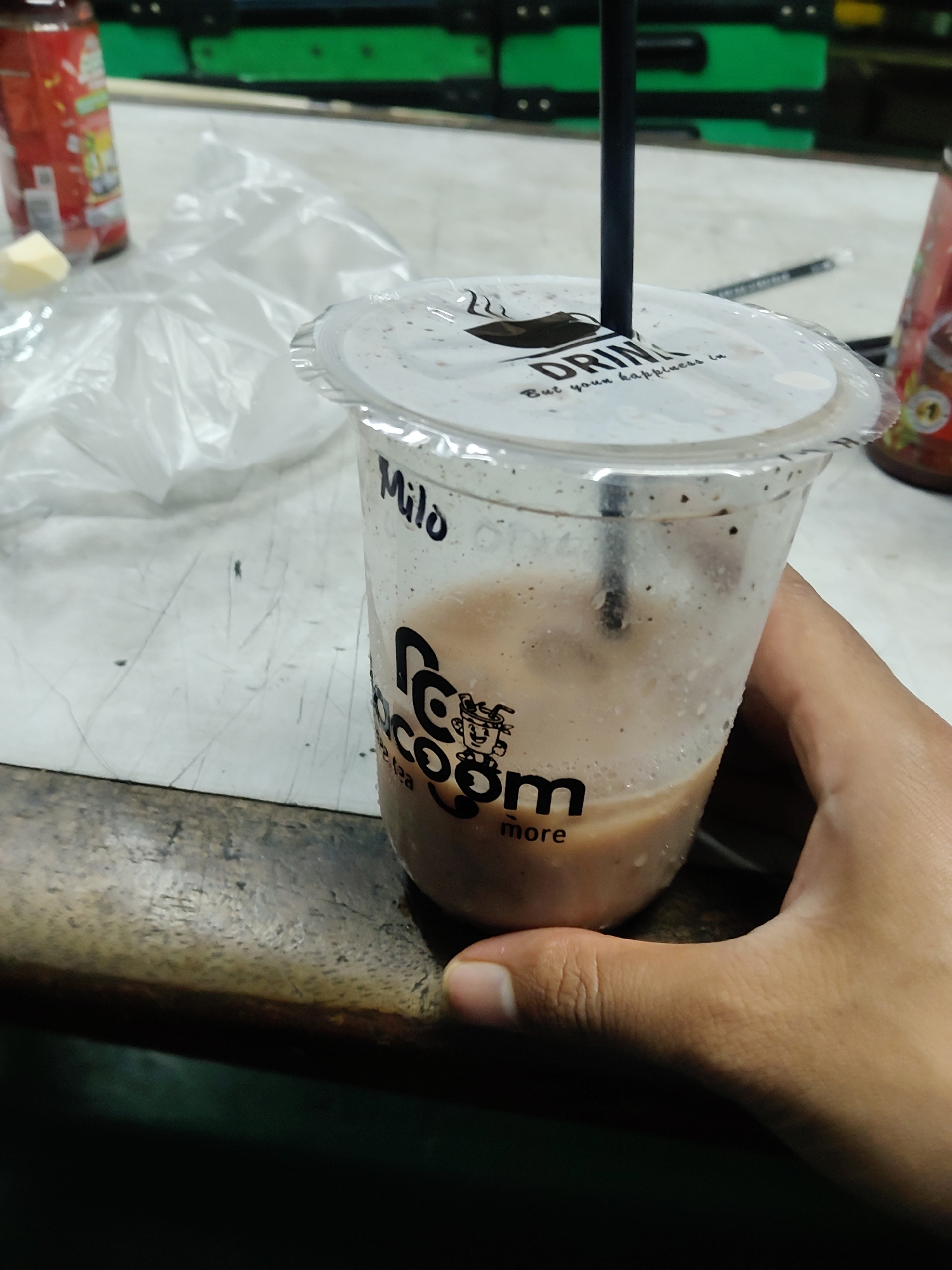 A close-up, eye-level shot captures a hand holding a clear plastic cup filled with a creamy brown beverage. The cup is partially filled, with a black straw inserted into a white lid that has a black logo and text reading "DRINK. But your happiness is". The cup itself has a black logo on it, a stylized character with "coccom" written vertically, and then "more" below. The words "Milo" and "ice tea" are also visible on the cup. The cup is sweating, indicating it contains a cold drink. In the background, a red bottle and a white plastic bag are visible on a wooden surface. The setting appears to be a casual eatery or street vendor stall in Mekarmukti, Indonesia, suggested by the presence of local branding and packaging often found in such locations. The lighting is somewhat dim, creating a slightly grainy texture to the image.