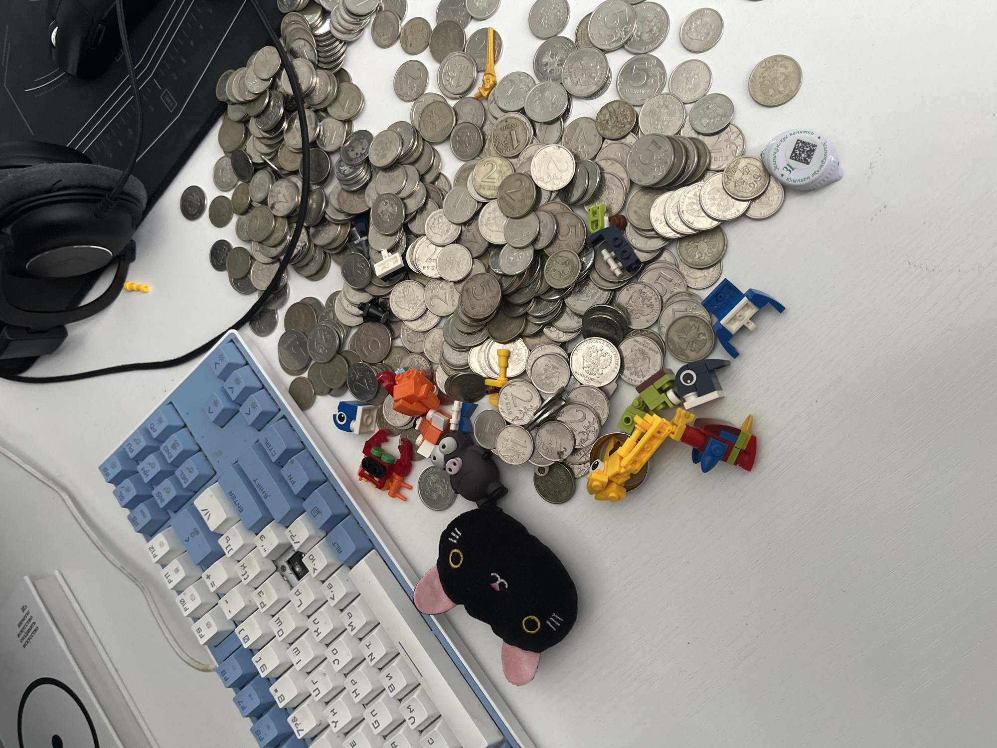 The image depicts a white, textured desk surface. Scattered across the desk are several objects including a light blue and white mechanical keyboard, a pile of silver coins, and various small plastic building toy pieces. A black plush toy in the shape of a cat's head with pink ears and yellow eyes rests near the keyboard. A pair of black headphones is visible in the upper right corner, alongside a black mousepad with a white grid pattern and a black computer mouse. The lighting suggests an indoor setting, likely during the day. No specific location in Saratov, Russia is identifiable.