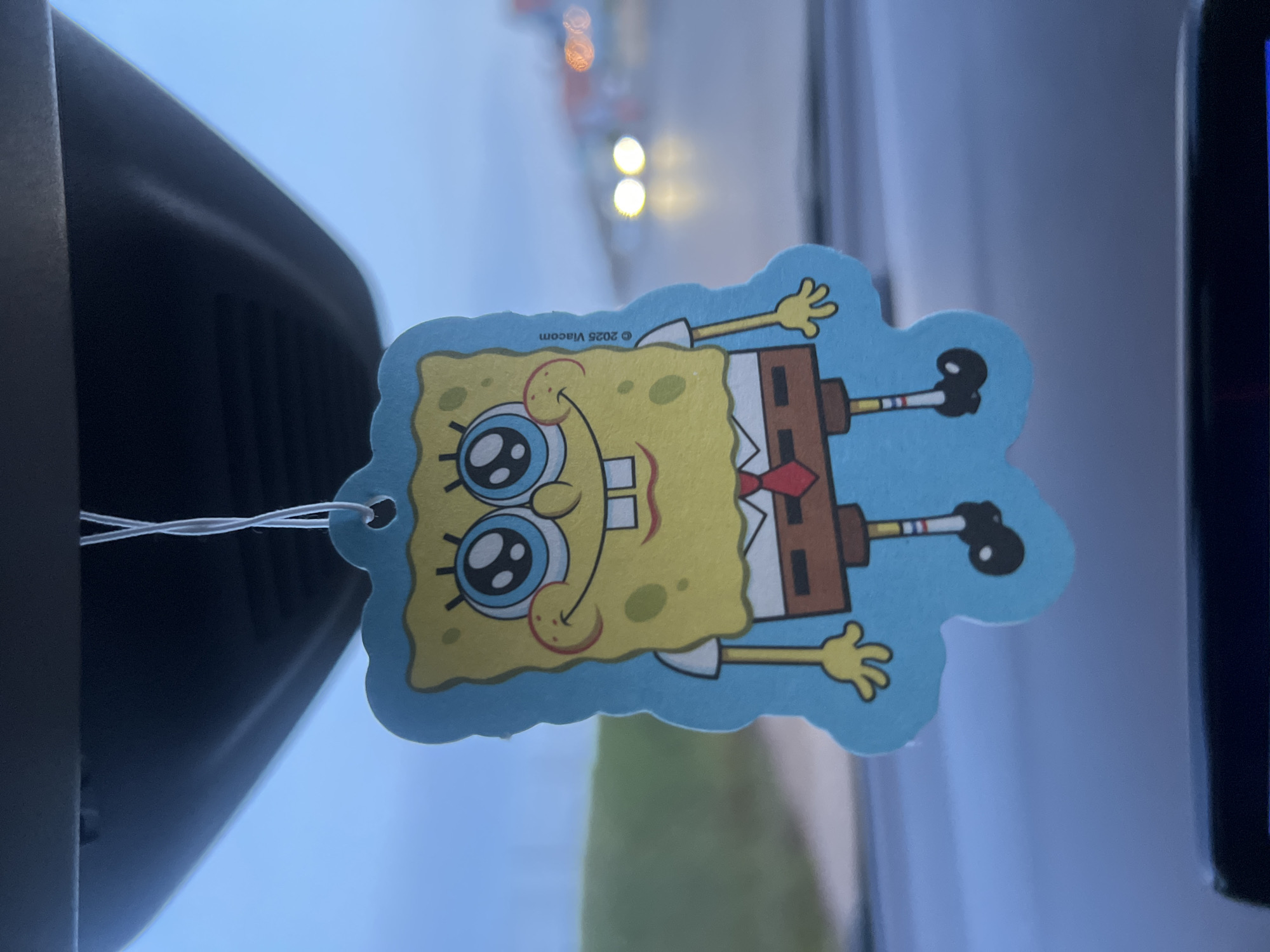 A SpongeBob SquarePants air freshener hangs from the rearview mirror of a car. The location is not discernible from the image. The environment appears to be indoors, possibly inside the vehicle, with a blurred background visible through the windshield. The car's interior trim is visible to the left of the air freshener.