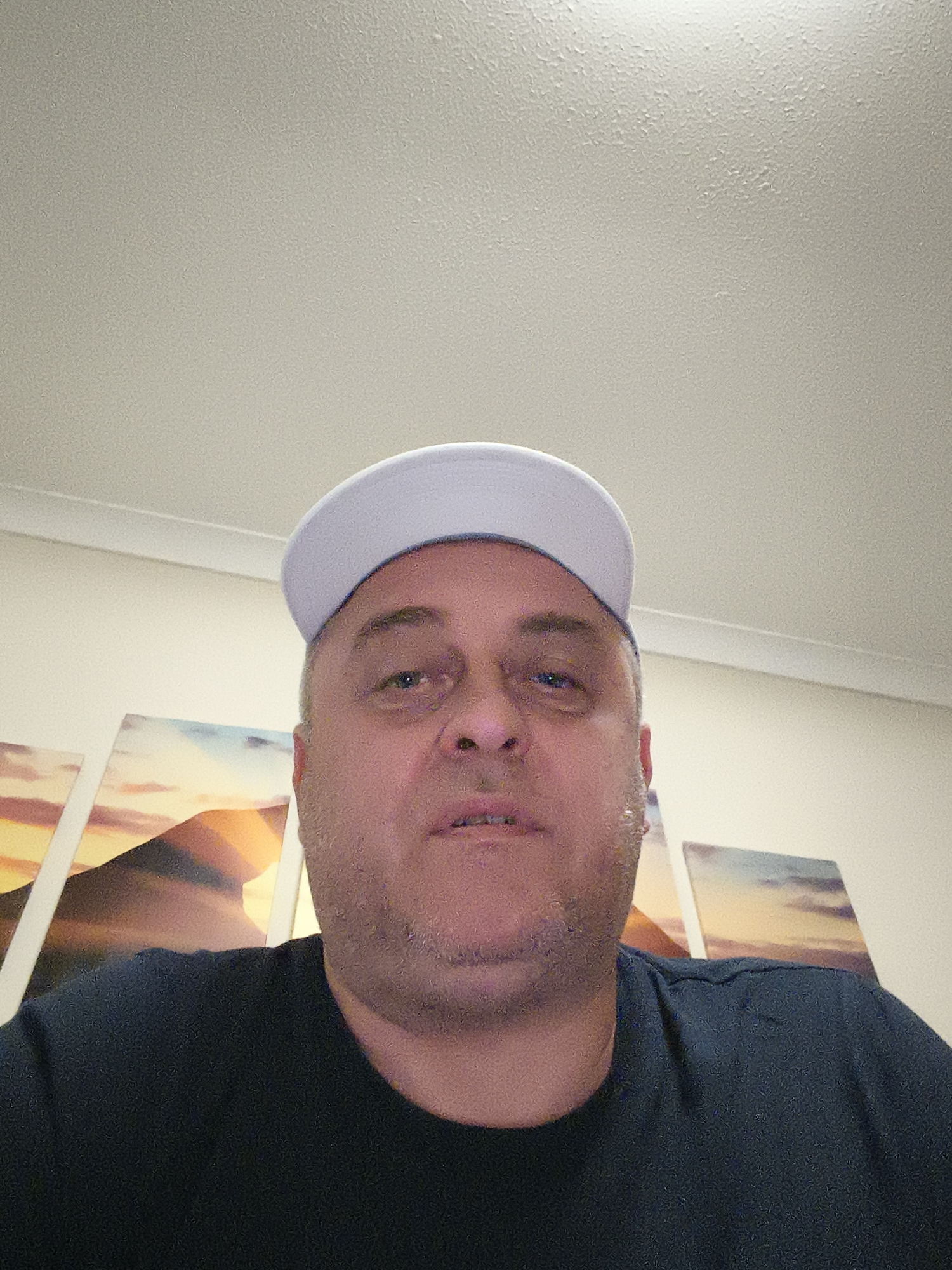 A man, appearing to be middle-aged, is the central subject of an indoor, chest-up portrait, likely a selfie. He faces the camera directly, exhibiting a neutral expression with slightly tired-looking, light-colored eyes. He has short hair and noticeable stubble on his face. He is wearing a white or very light grey baseball cap, positioned with the brim forward, and a dark-colored, short-sleeved t-shirt. Artificial overhead lighting illuminates his face and the textured details of his skin and attire.
The background reveals a domestic or office setting. On the wall behind the man, multiple pieces of artwork are displayed horizontally. These appear to be canvases or framed prints, showcasing a serene landscape with warm hues of orange, yellow, and blue, possibly depicting a sunrise or sunset over a natural formation. Above the man's head, a distinctly textured white ceiling is visible, meeting the wall at a clean, white molding. No outdoor elements or specific location cues beyond an indoor environment are discernible, though the general style is consistent with a residence in the Huntingdonshire, United Kingdom area. There is no visible text in the image.