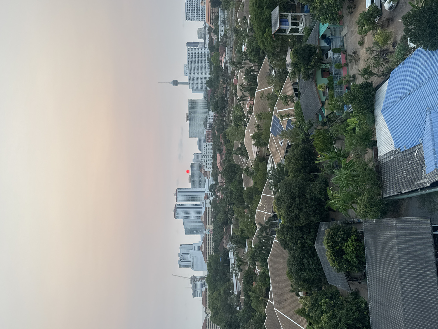 This high-angle, panoramic view captures a diverse landscape in Pattaya City, Thailand, likely during sunrise or sunset. The horizon line stretches across the upper middle of the frame, revealing a prominent city skyline dominated by numerous modern high-rise buildings and condominiums. A distinctive observation or communication tower is visible among the distant structures, a recognizable landmark of Pattaya. A small, reddish-orange orb, indicating the sun, is visible in the hazy sky near the horizon, casting a soft, warm glow that fades into a lighter grey.
Below the city skyline, the midground and foreground are densely covered with lush tropical vegetation, including a variety of trees and palm trees, interspersed with residential and perhaps some commercial buildings. Many of these structures feature brown-tiled roofs, and several are equipped with solar panels. In the immediate foreground, a range of roof types is visible, including prominent blue corrugated metal roofs and darker grey, flatter roofs. The image presents a blend of urban development and natural greenery under soft, ambient light, with no visible people or text.