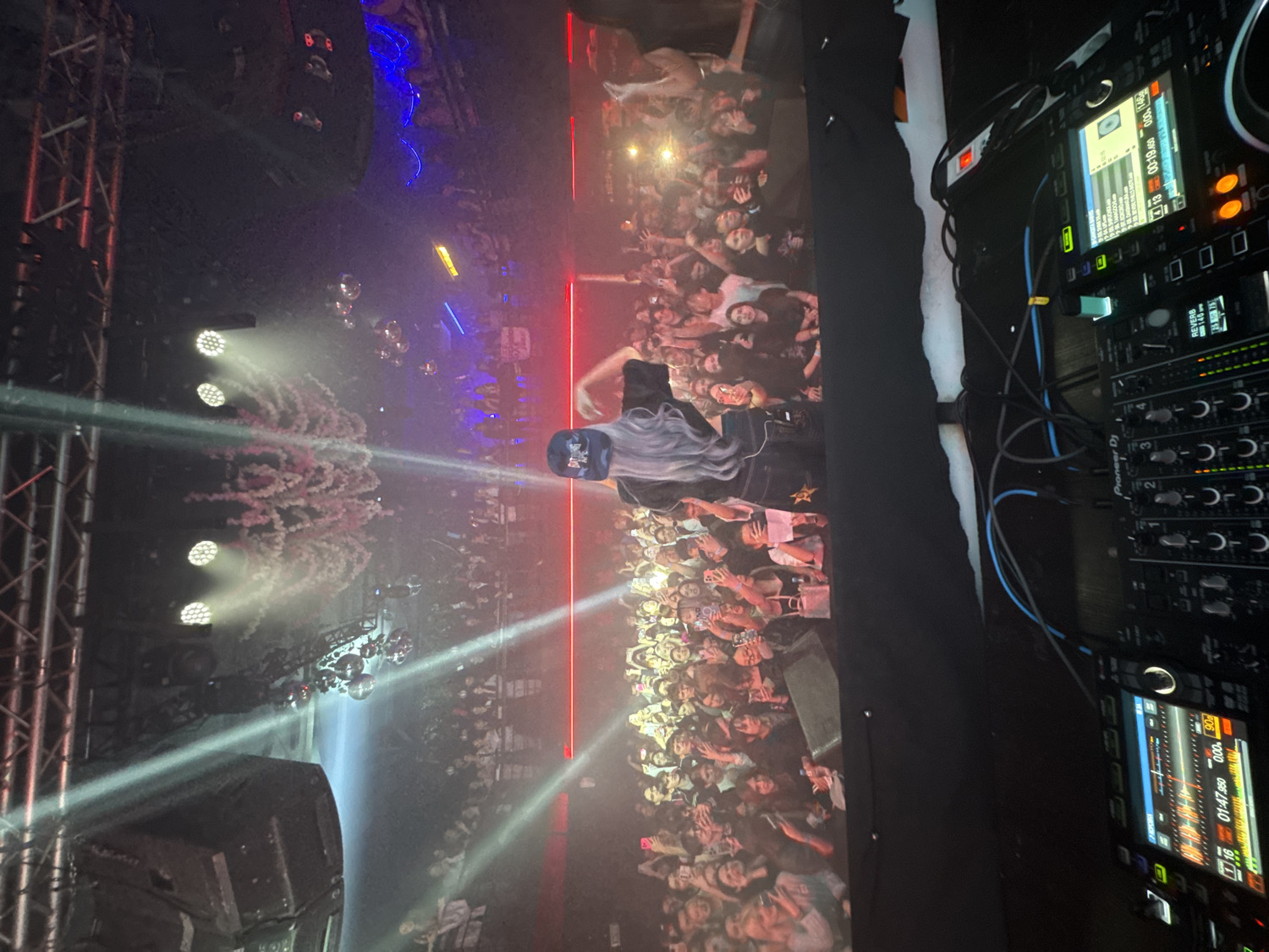 This image captures a vibrant night concert or club event, taken from the perspective of a DJ booth looking out towards a stage and audience. The scene is illuminated by dynamic stage lighting, creating a high-energy atmosphere.
On the right side of the frame, partially visible, is a DJ, seen from behind, with long, light-colored hair, positioned behind a professional mixing console. The console prominently features "Pioneer Dj" branding and several control knobs labeled "C-C-". Its screens display digital information, including track times like "01:47.550" and "00:19.450", and the word "REVERB". Cables are visible connecting different parts of the equipment.
In the center and left, a large crowd of people is densely packed, facing the stage and performer. Many in the audience are looking up, some with their hands raised, indicating engagement and excitement. The stage area features bright, white spotlights emanating from truss structures on the far left. Above the crowd, multiple disco balls reflect light, adding to the festive mood. Pink and white confetti or pyrotechnic smoke effects are seen billowing upwards from the stage area, enhancing the visual spectacle. Red and blue ambient lights cast a glow over parts of the venue, while strong light beams cut through the smoky air. A large speaker is visible in the lower left corner.
The overall impression is one of an energetic and lively indoor event, likely a music performance, taking place at night in a venue in Vladivostok, Russia.