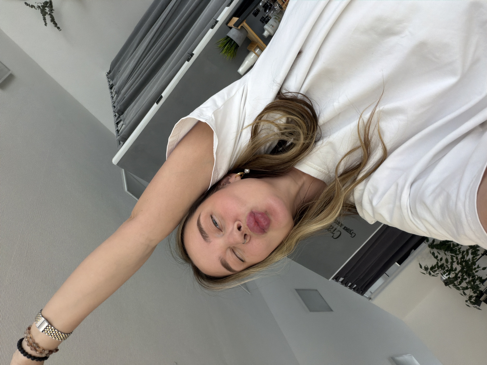 A young woman with blonde hair is making a playful face. She is wearing a white T-shirt and has jewelry on her wrist and an earring. The background shows part of a room with gray curtains, a plant, and some decorative items. Based on the text partially visible on the wall, the location may be Chaikovsky, Russia.