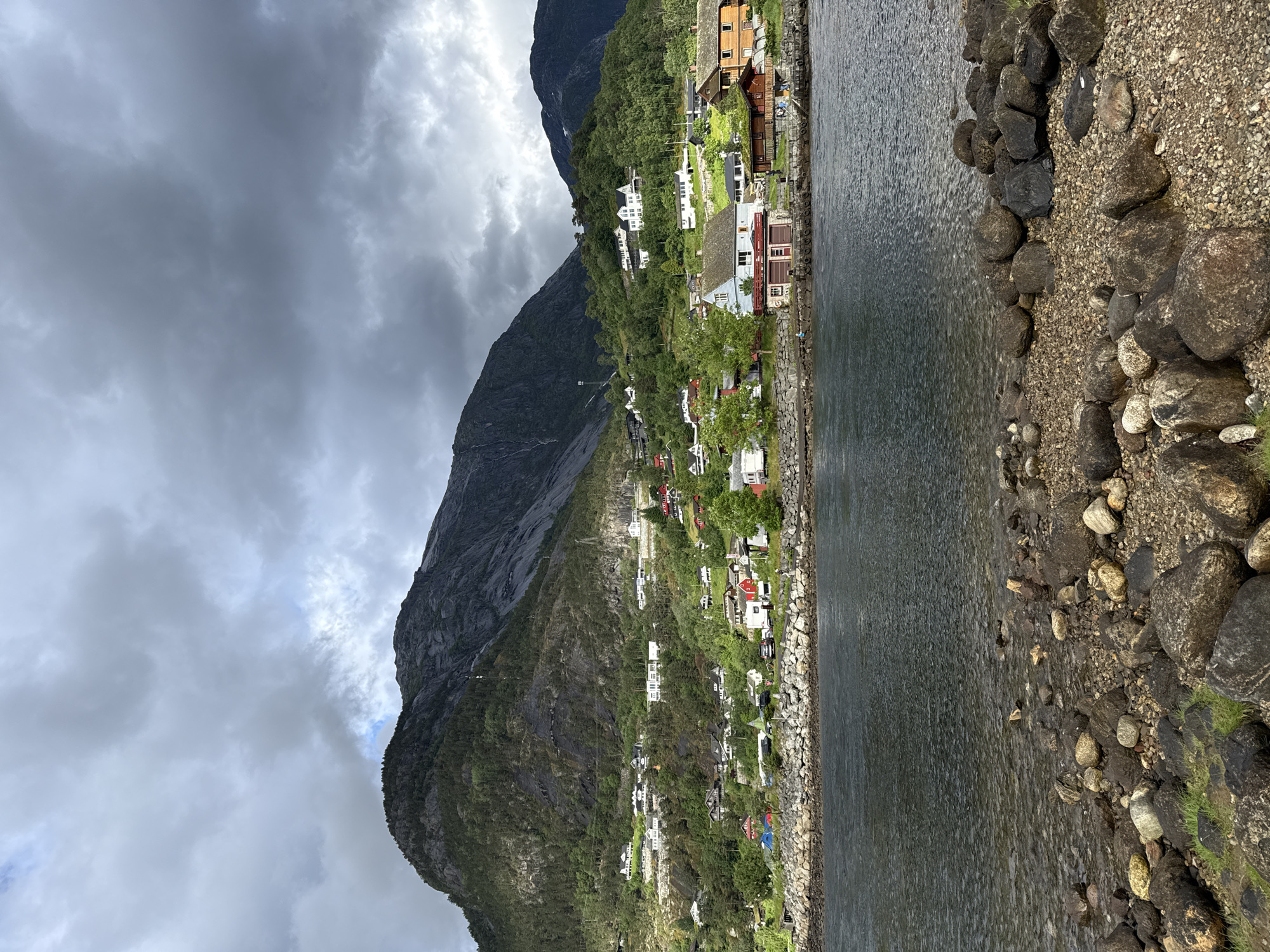 A scenic view of Eidfjord, Norway, showcases a serene body of water in the foreground, with a rocky shore dotted with pebbles and grass. The water reflects the overcast sky, creating a subdued palette. Across the water, a charming village is nestled on a lush green hillside, with houses of various colors and architectural styles. The village extends up the slope, with some homes perched higher on the hill. A majestic mountain range dominates the background, its slopes covered in dense evergreen trees. The mountain's rugged terrain is partly obscured by dramatic, dark storm clouds, suggesting an impending change in weather. The overall scene evokes a sense of tranquility and natural beauty, characteristic of the Norwegian landscape.