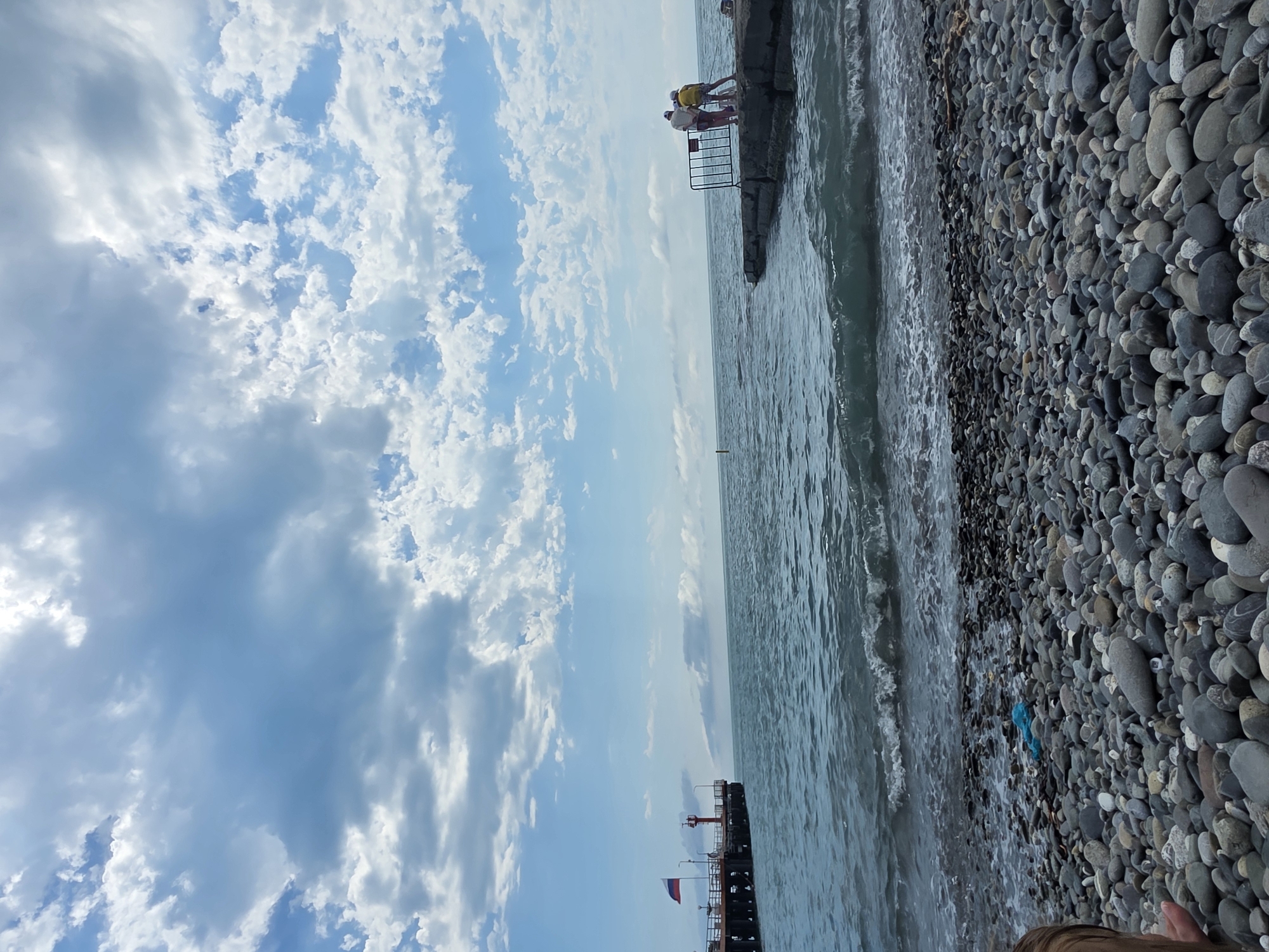 The image depicts a view of the Black Sea coast in Sochi, Russia. In the foreground, a pebble beach is visible, with scattered stones of various sizes and shades of gray. A piece of blue plastic is lying on the beach. Waves are gently lapping at the shore.
In the mid-ground, the sea stretches out towards the horizon under a partly cloudy sky. The water appears choppy with whitecaps. To the left, a pier extends into the sea, featuring a red lighthouse and a Russian flag flying from a pole.
In the right mid-ground, a concrete jetty juts out into the water. Two individuals, one wearing a yellow shirt and the other a beige shirt, stand on the jetty, looking out at the sea. A black metal railing is positioned behind them. The sky is a mix of blue and white clouds, with some areas of brighter light suggesting sunshine breaking through.
