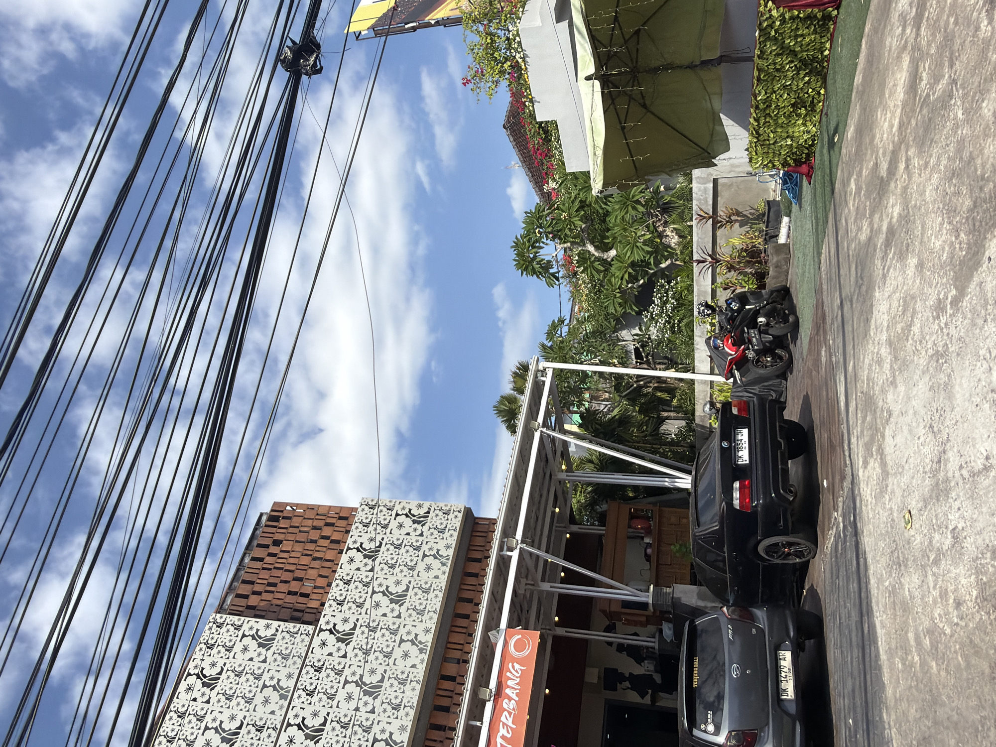 This image shows a street scene in Kuta, Indonesia, on a bright, sunny day with some clouds. The perspective is tilted, looking upwards at a cluster of utility wires stretching across the frame. Below the wires, there is a building with a patterned facade and a red-brick upper section. Adjacent to this building is an outdoor dining or seating area with a white canopy and tables.
In front of the building, two cars are parked on a paved surface. The car closest to the viewer is a gray hatchback with license plate "DK 1479 AR". The car behind it is a black sedan with license plate "DK 1854 JH". A motorcycle is also parked in the scene.
Green foliage and flowering plants are visible, suggesting a tropical environment. A large umbrella, likely for shade, is partially visible at the top. A banner with the word "TERBANG" is hanging from the building. Another sign reads "TUNJUNG BALI". The overall impression is of a lively commercial area, possibly a restaurant or cafe, on a typical day in Bali.