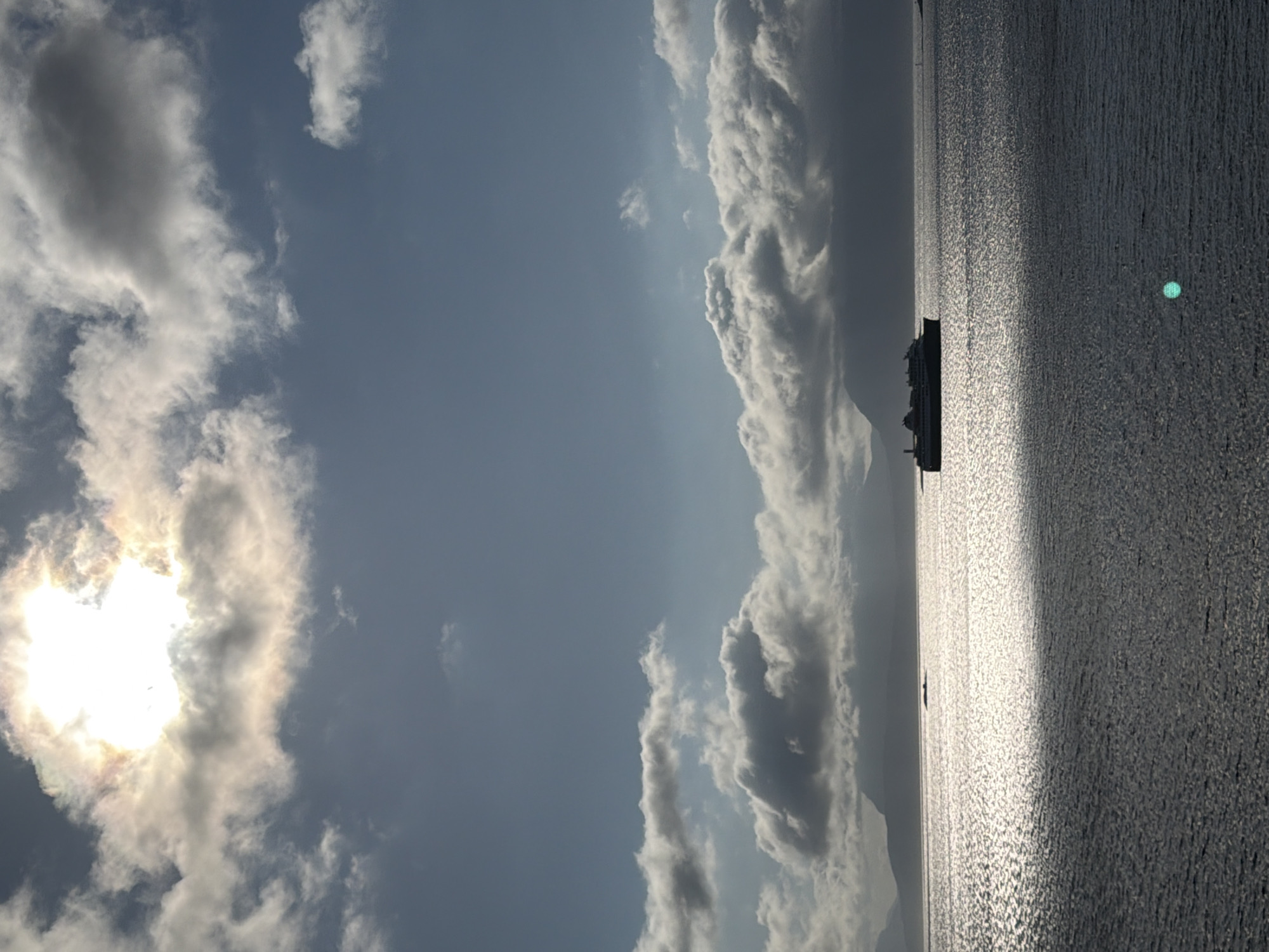 The image depicts a vast body of water, likely the Harstad harbor, under a dramatic sky. The sun is partially obscured by thick, white clouds, casting a bright, diffused light that reflects intensely off the rippling water. In the middle ground, a large ferry is silhouetted against the bright water, its dark shape starkly contrasting with the shimmering surface. In the background, the dark, rugged outlines of mountains or islands rise from the horizon, also silhouetted against the sky. The water is textured with numerous small waves, creating a shimmering, almost metallic appearance. A faint green circular glint, possibly a lens flare or reflection, is visible on the water's surface in the lower right quadrant. The overall mood is serene yet powerful, with the vastness of the water and sky dominating the scene.