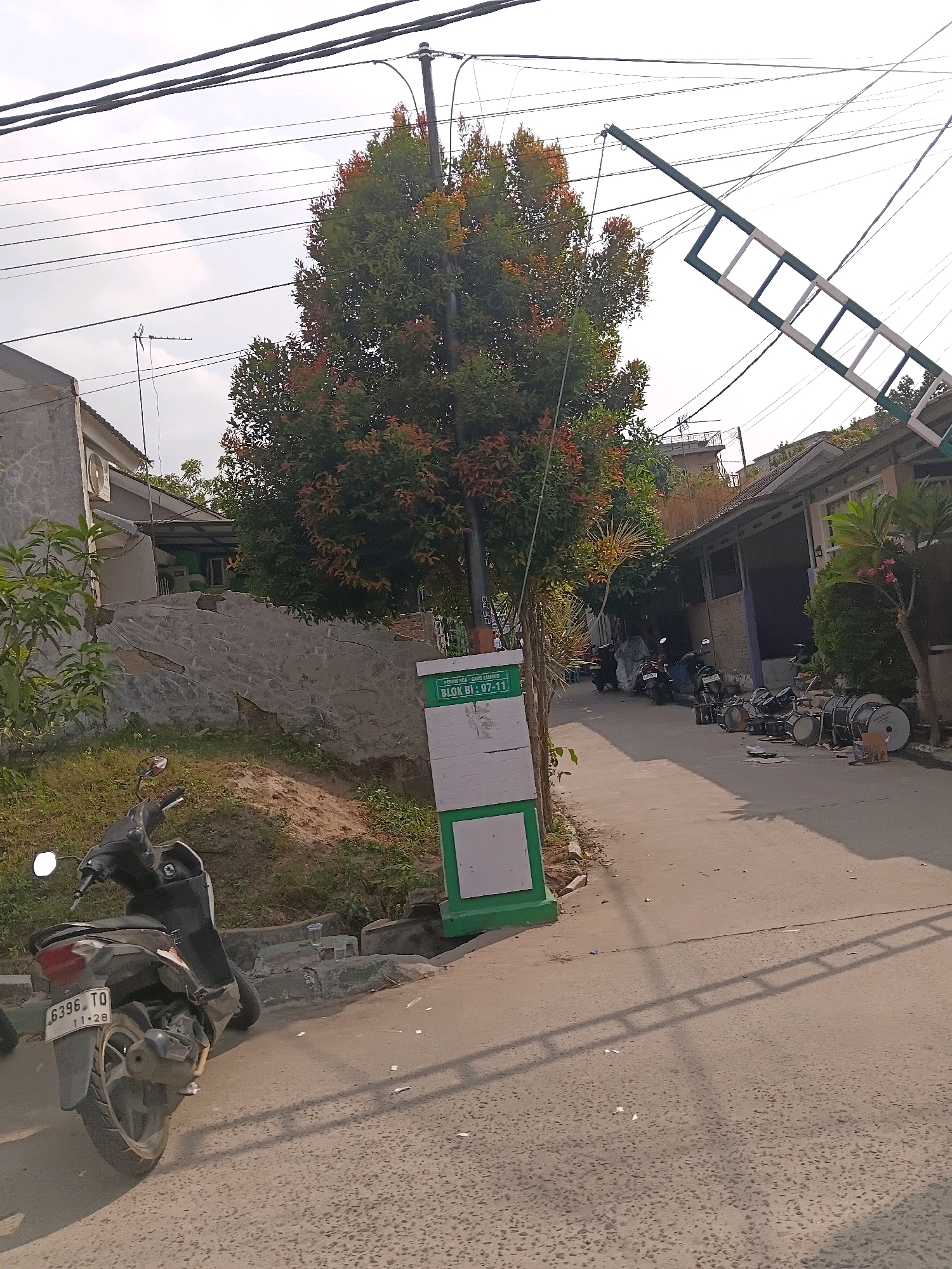 The image captures a street scene in Cilegon, Indonesia, likely a residential or commercial alleyway, under a partly cloudy sky. A large tree with reddish-green foliage stands prominently in the middle of the frame. To the left, a residential building with a grey stone wall and an air conditioning unit is visible. A dark-colored motorcycle with license plate "B396 TO 11-28" is parked on the left side of the paved street. A tall, green and white signpost with text that appears to be in Indonesian is located near the tree.
The street curves to the right, revealing several parked motorcycles and what appear to be large drums or percussion instruments arranged on the right side of the road. The buildings on the right have pitched roofs and some have open-fronted structures. Power lines crisscross overhead. A long, green and white structure resembling a ladder or ramp is suspended diagonally across the upper right portion of the image, attached to a pole. Sunlight casts shadows of the power lines and the suspended structure onto the street, indicating it is daytime. The overall impression is of a somewhat cluttered but lived-in urban environment.