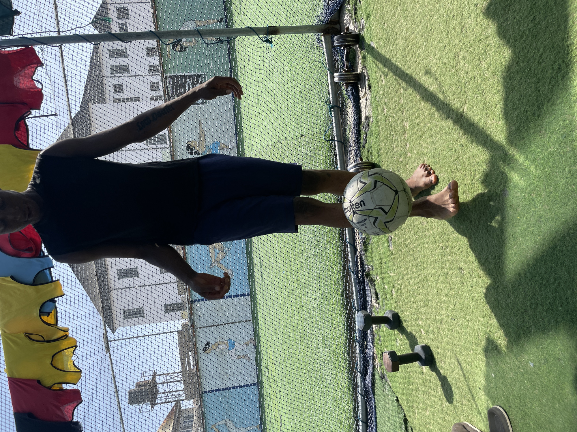 The image shows a person demonstrating soccer skills on an outdoor artificial turf field. The subject, an individual with dark skin, is dressed in dark shorts and a black sleeveless top. They are balancing a white and green "molten" branded soccer ball on their left foot/ankle, with arms slightly raised for balance and concentration.
The setting is a vibrant green artificial grass pitch, characteristic of a sports training ground. Strong shadows cast by the person, a pole, and the netting suggest bright daylight, indicating clear and sunny weather, likely midday or early afternoon. To the left of the person, a stack of colorful training vests (red, yellow, blue) is visible, hanging on a pole. Two dumbbells rest on the grass to the right. A protective net separates the immediate foreground from the background, which features multi-story buildings under a clear sky.
A tattoo on the person's left forearm is partially visible, appearing to read "Legal Death". The scene conveys a focused moment of athletic practice within an urban or suburban sports facility, consistent with locations like Iranla, Nigeria.