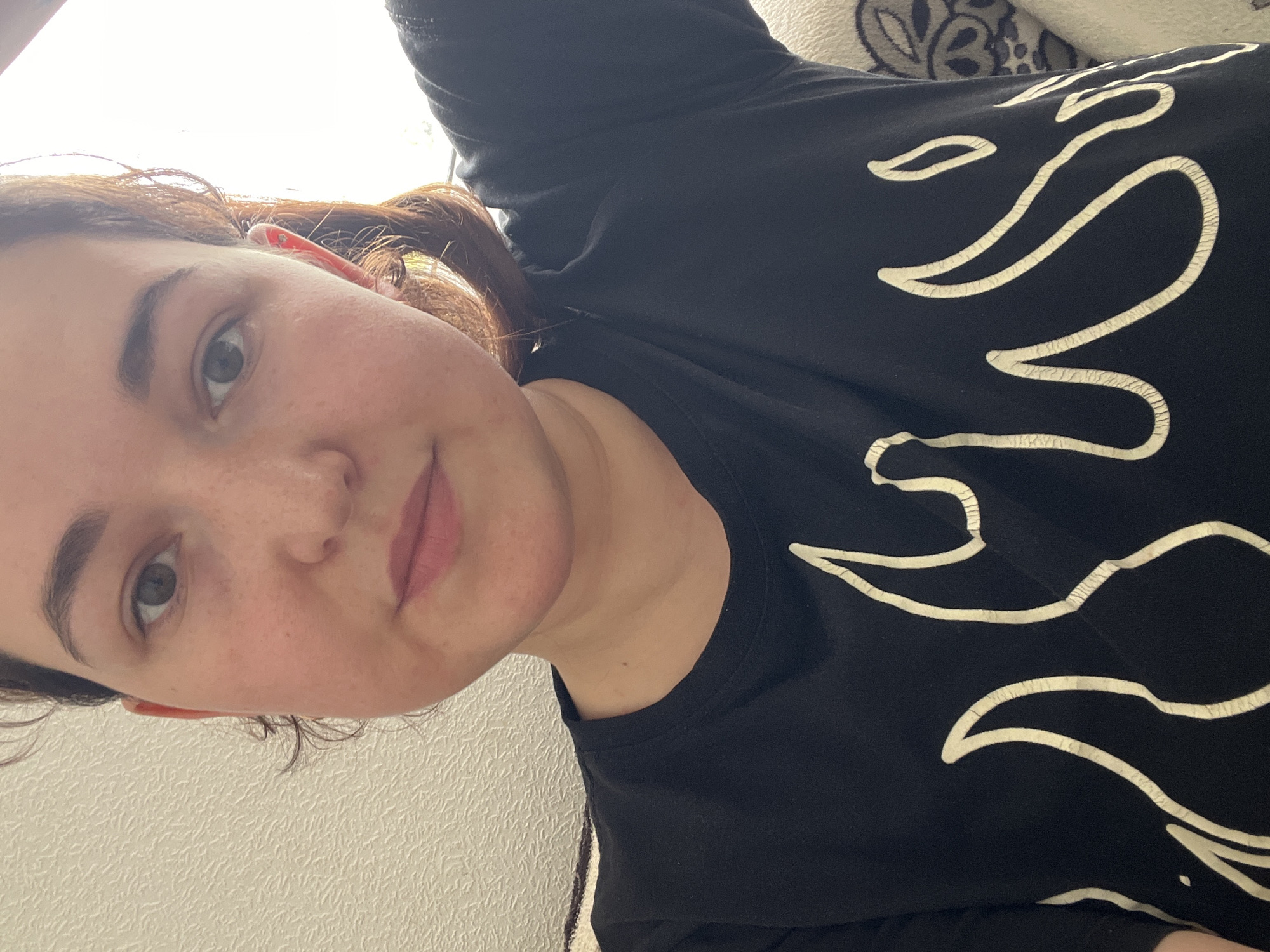 The image shows a close-up, chest-up selfie of a young woman. She has fair skin with freckles, light brown hair pulled back, and is wearing a black t-shirt with a white flame design. The background is blurred and appears to be indoors, with a textured wall on the left and bright light coming from the right, possibly a window. No specific location in Chelyabinsk, Russia can be identified.
