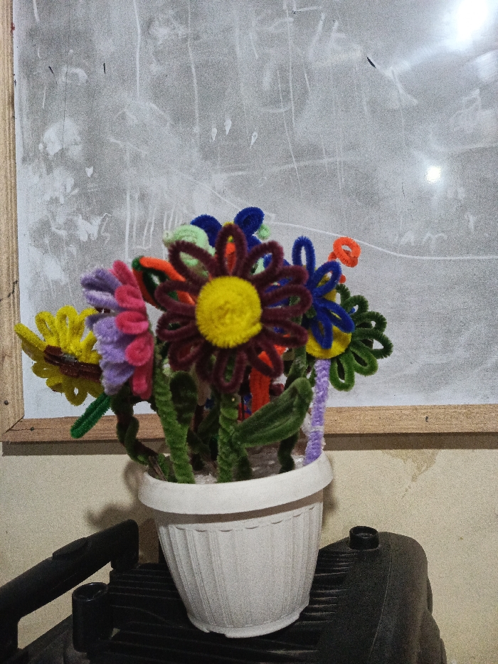 The image displays a vibrant bouquet of artificial flowers, crafted from colorful pipe cleaners, set in a white, ribbed plastic pot. The flowers feature an array of colors including yellow, purple, pink, maroon, orange, blue, and green, each centered with a fluffy yellow pom-pom. Green pipe cleaners form the stems and rudimentary leaves, some twisted to add texture.
The white pot is placed on a dark, ridged surface, possibly part of a piece of equipment or furniture. Another dark, indistinct object is partially visible on the left side, beneath the pot. The background consists of a light gray surface, resembling a whiteboard or a painted wall, showing numerous white scuff marks, smudges, and faint, indistinct white markings that might be remnants of past writing or drawings. A light brown wooden frame borders this background surface on the left and bottom edges.
The scene is an indoor still life, characterized by dim lighting, suggesting either evening or a poorly lit room. There are no people or activities depicted. The handcrafted flowers convey a sense of simple, cheerful artistry. No clear or legible text is visible in the image. The setting appears to be a casual environment.