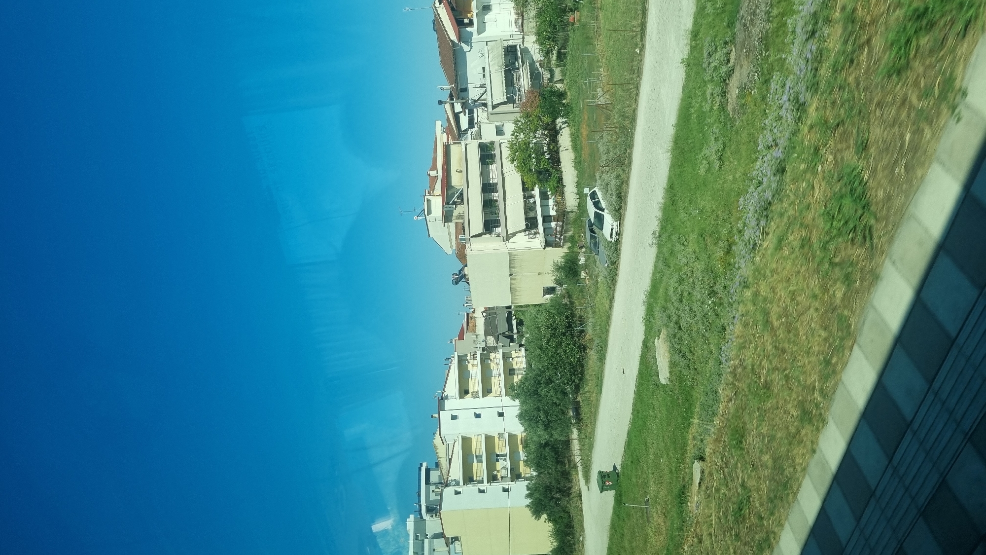 The image captures a view of residential buildings under a clear blue sky, likely from a moving vehicle given the slight blur in the foreground. The buildings are multi-story, painted in light colors such as yellow and white, with balconies. Greenery, including trees and grass, surrounds the buildings. A dirt road runs in front of the buildings, with two cars parked on it and a green trash bin to the left. The scene appears to be in a suburban or semi-urban area. The presence of the specific architectural style of the buildings and the characteristic Mediterranean vegetation suggest a location in Greece. Specifically, the image is consistent with a scene in the city of Larissa, Greece, which has similar residential areas and landscapes.