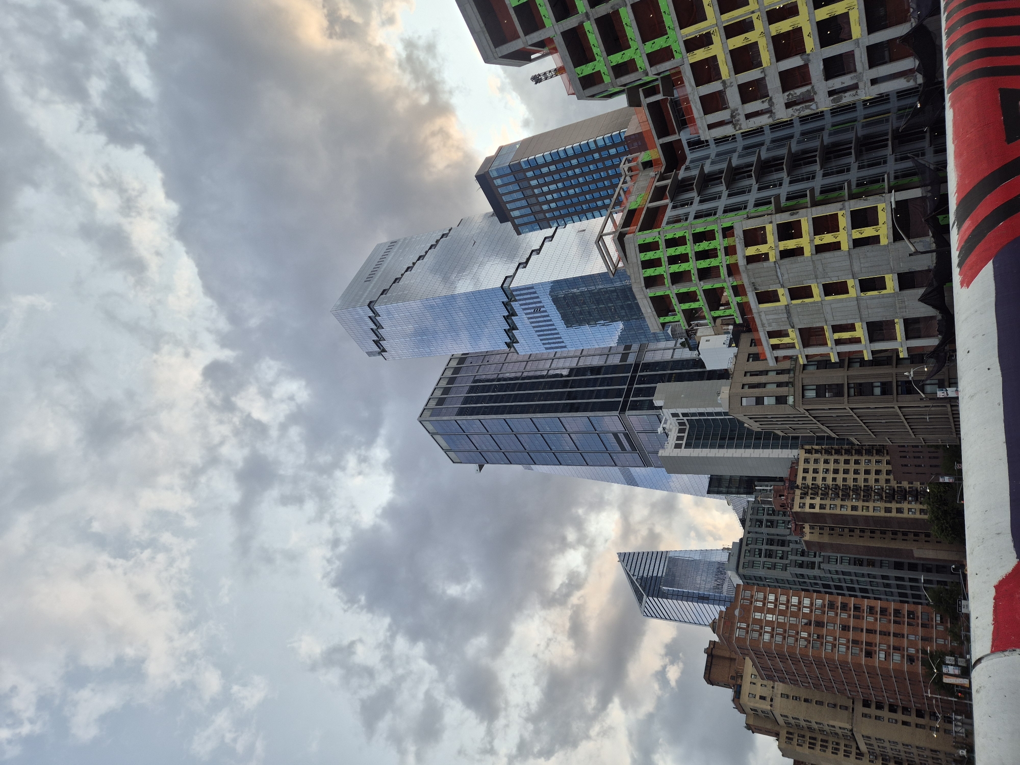 This is a low-angle, wide shot of a New York City skyline dominated by various skyscrapers and buildings under construction. The sky is filled with dramatic, heavy gray clouds, with hints of golden light breaking through on the right side. In the foreground, a white concrete barrier with red and black stripes runs across the bottom of the frame.
Several distinct buildings are visible. The most prominent is a modern, glass-faceted skyscraper with a distinctive stepped design and shimmering blue glass facade, reflecting the sky. To its left, another tall, sleek glass skyscraper with a triangular top is visible. Further to the right, a darker, rectangular skyscraper with many windows stands tall. In front of these completed structures, several buildings are in various stages of construction, their concrete frameworks exposed and covered with green and yellow sheathing. Some have orange netting draped around them. Older, brick and tan apartment buildings are visible on the left side of the frame, offering a contrast to the modern architecture. Streetlights and trees are sparsely visible at the base of the buildings. The image appears to have been taken from an elevated position, possibly a bridge or overpass, looking up at the cityscape. The overall impression is one of urban dynamism, with ongoing development alongside established architecture under a moody sky.
