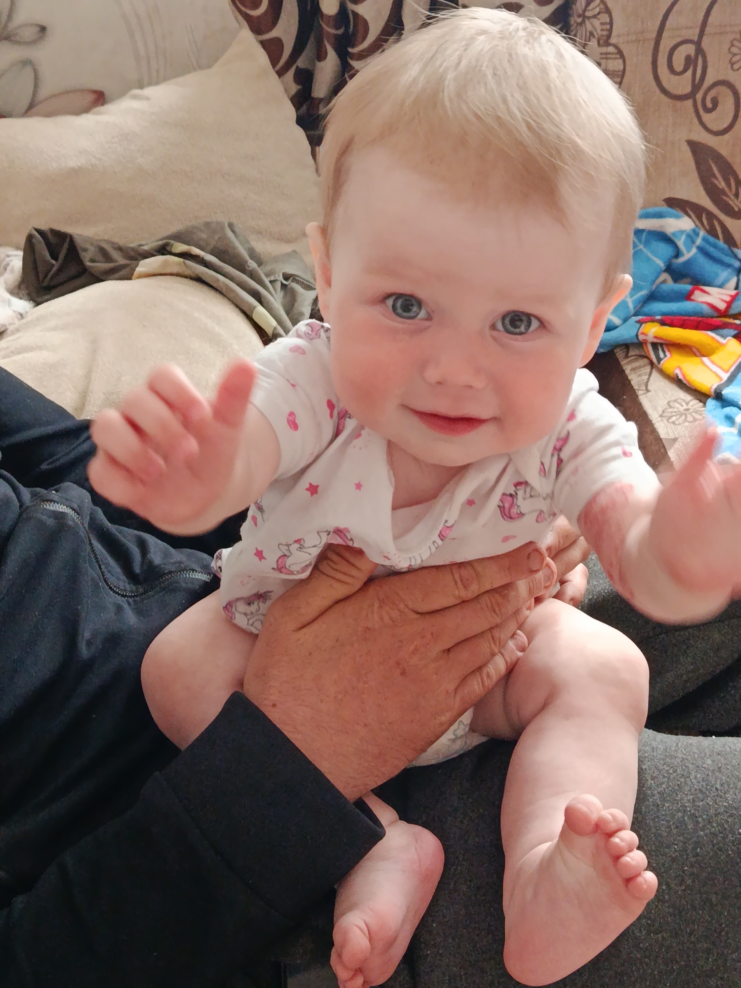A fair-skinned baby with light blonde hair and blue eyes sits in an adult's lap. The baby wears a white onesie with pink unicorn and star patterns. The adult's hand is visible, holding the baby securely. The adult wears a black long-sleeved shirt and gray pants. The background includes a beige pillow, a patterned brown and beige couch, and a blue blanket with cartoon imagery. Location: фоки, Russia (deduced based on the user profile).