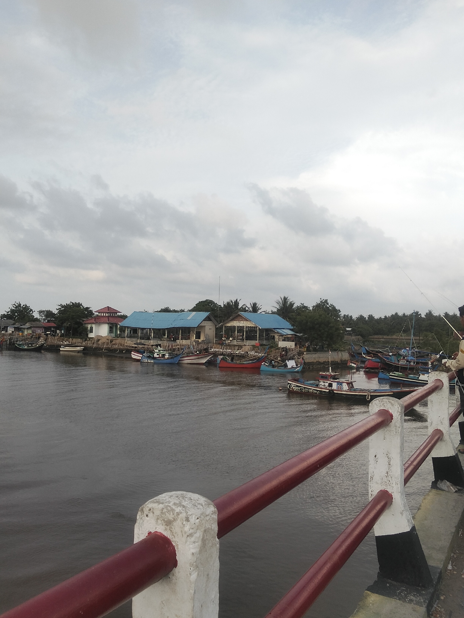 This is a wide-angle, eye-level photograph taken in Tambonbaroh, Indonesia. The scene depicts a waterfront with a river lined by buildings and numerous fishing boats. In the foreground, a red and white railing separates the viewer from the murky, rippling water. Several boats, mostly of traditional Indonesian design, are moored along the bank. Some buildings with blue and red roofs are visible, interspersed with lush green trees. The sky is overcast with gray clouds. On the right side of the image, a person is standing on the embankment, holding a fishing rod. The overall atmosphere is that of a working fishing village on a cloudy day.
