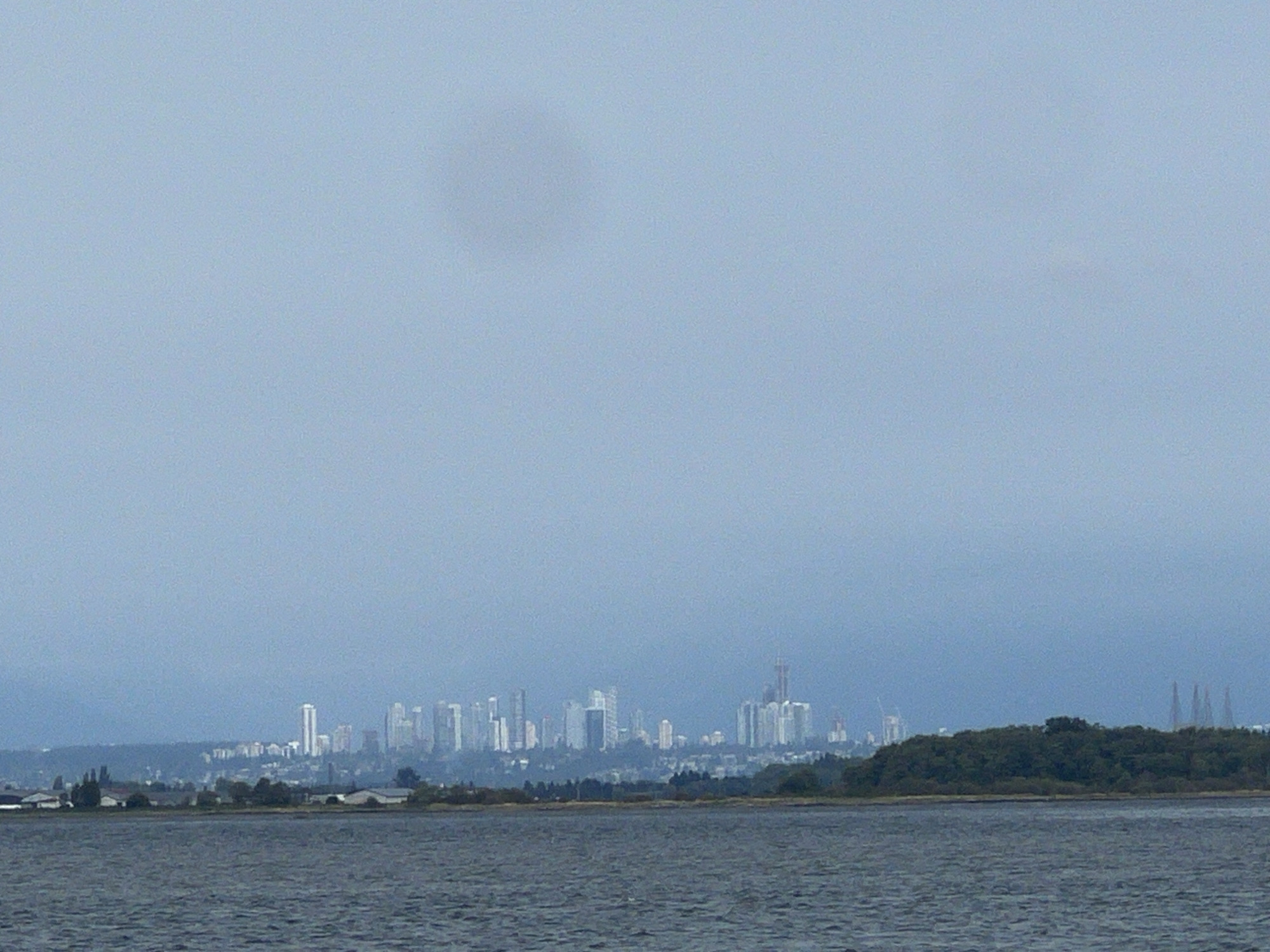 The image depicts a hazy, distant view of the Vancouver skyline from across the Fraser River, likely taken from the city of Surrey, Canada. In the foreground, a large body of dark, rippling water occupies the bottom portion of the frame. Beyond the water, a low-lying, tree-covered shoreline is visible, with a few structures, possibly industrial buildings or warehouses, situated near the water's edge. Further inland, the densely packed skyline of Vancouver rises, characterized by numerous high-rise buildings of varying heights and designs. The sky is overcast and a pale blue, contributing to the muted and somewhat indistinct appearance of the distant city. The overall scene conveys a sense of urban sprawl viewed from a distance, with the natural element of the river and shoreline providing a contrast to the man-made structures of the city.