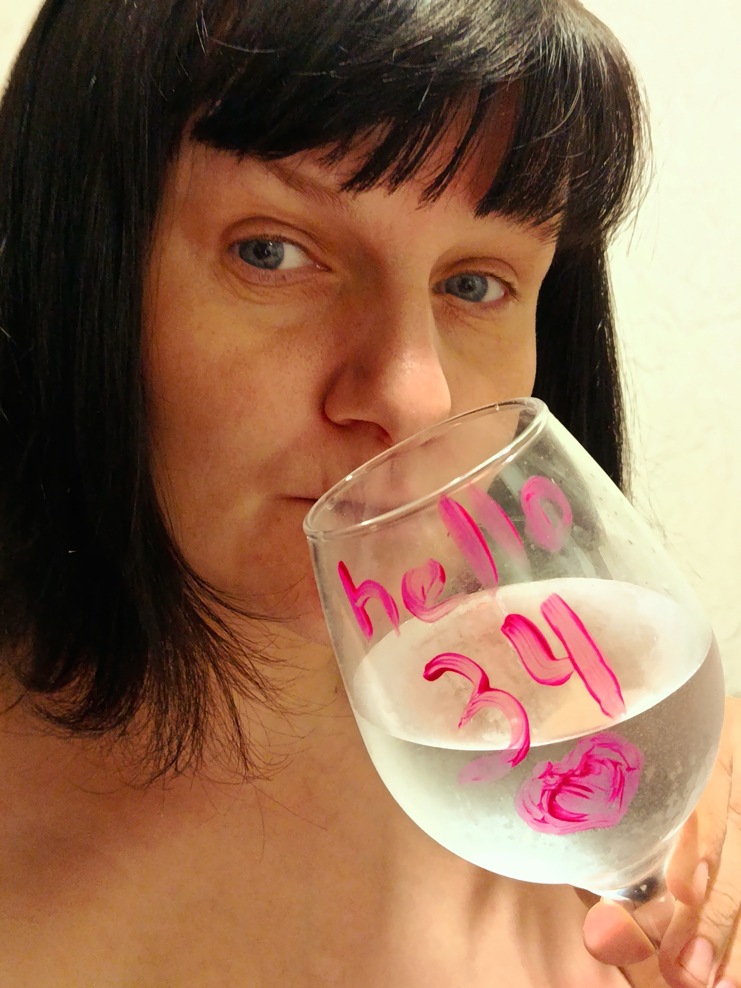 A close-up portrait shows a person, likely a woman, with dark hair and bangs, looking directly at the viewer with blue eyes and a neutral expression. She holds a large, clear stemmed glass, tilted, from which she appears to be drinking or about to drink. The glass contains a clear liquid. Written on the glass in bold pink script are the words "hello" and the number "34," followed by a pink heart design. This text suggests a birthday celebration or an acknowledgement of turning 34 years old. The background is a plain, light-colored wall, indicating an indoor setting. No specific time of day or location cues are visible within the image to suggest its setting in Фоки, Russia, beyond the context provided. The focus is on the individual and the celebratory message on the glass.