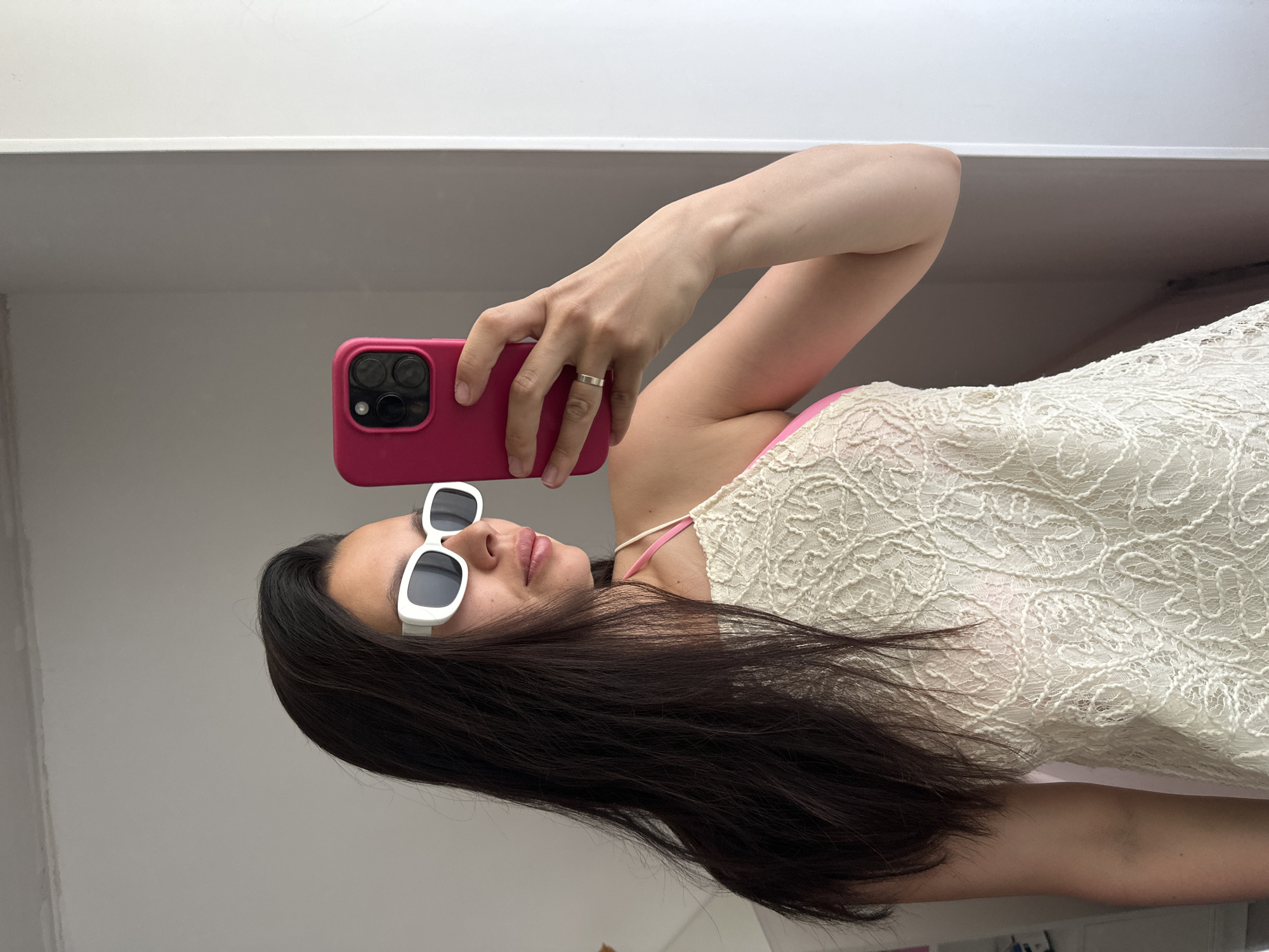 A woman is taking a selfie indoors. She is wearing white sunglasses, a white lacy top over a pink tank top, and is holding a pink phone. She has long dark hair. The location appears to be an interior space with white walls. The city is unspecified.