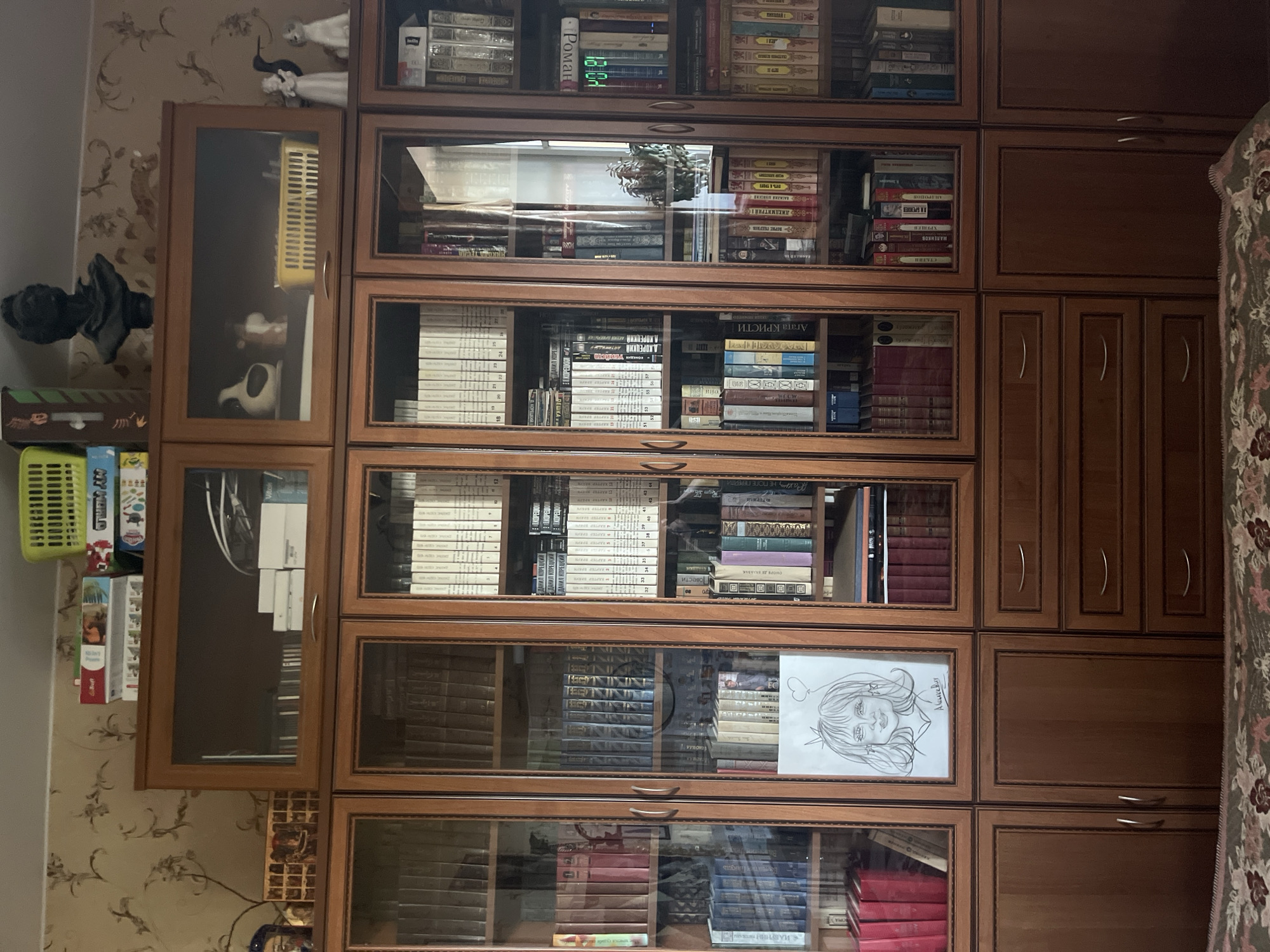 A full shot shows a wooden bookcase filled with books and various decorative items. The bookcase is situated in a room with patterned wallpaper. On the top shelf of the bookcase, there are stacks of board games, a green basket, and a black bust. The shelves are filled with books of various sizes and colors. Some shelves also hold decorative objects, including figurines and a framed drawing of a woman. The lower part of the bookcase has wooden cabinets and drawers. The scene is indoors, and the lighting appears to be natural, possibly from a window. The overall impression is one of a well-decorated and lived-in space. This image is located in the city of Vladimirovka, Russia.