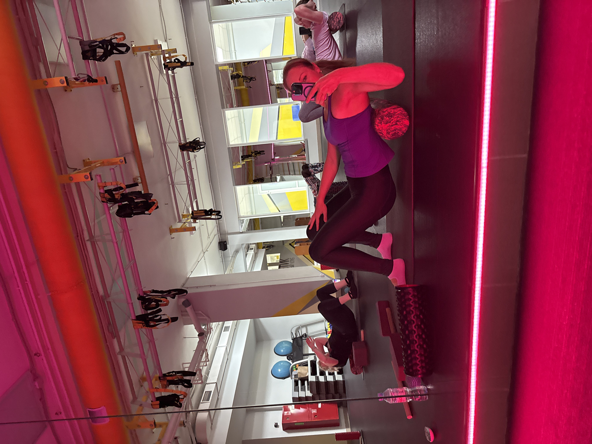 The image shows the interior of what appears to be a fitness studio or gym in Chaikovsky, Russia, as indicated by the Russian text visible in the background.
**People:**
- A woman is taking a selfie with a smartphone. She is wearing a purple tank top and dark leggings. Her expression is neutral.
- Another person is lying on the floor, possibly using a foam roller.
- There is at least one more person in the background engaged in some form of workout activity.
**Objects:**
- Foam rollers of different colors.
- Bottles of water.
- Exercise equipment hanging from the ceiling, arranged in rows.
- A mirror.
- Red fire extinguisher.
**Actions:**
- The woman is taking a selfie.
- Other people are exercising.
**Environment:**
- A brightly lit fitness studio.
- The color scheme includes yellow, gray, and pink.
- The space seems modern and well-equipped.