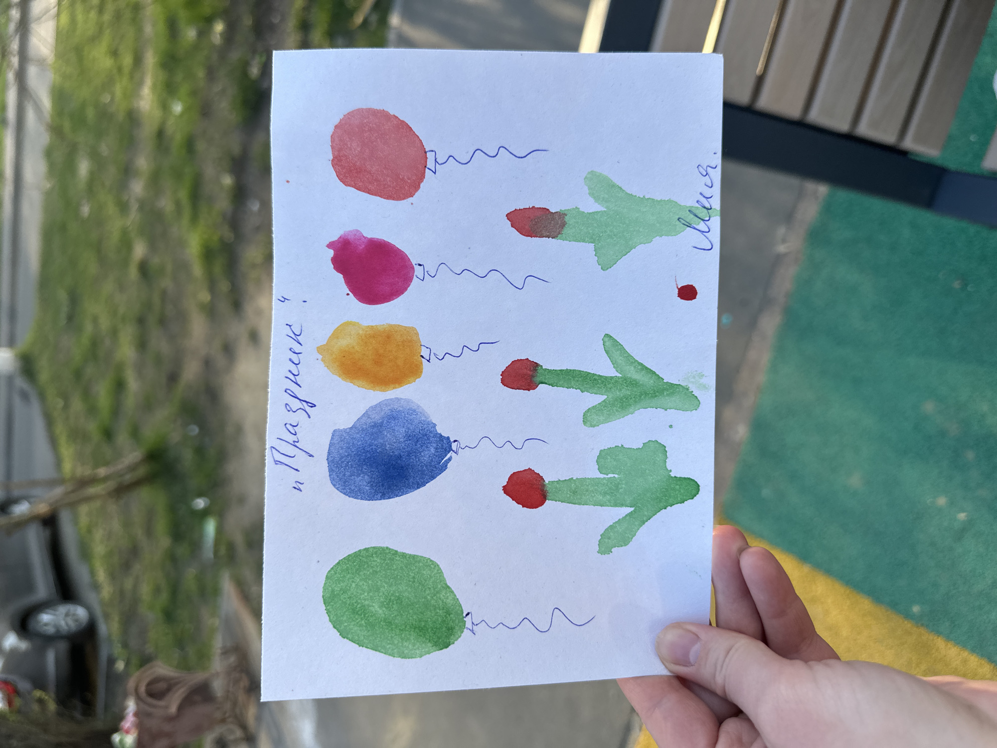 A hand holds a child's watercolor painting on white paper. The painting depicts five balloons on the left, rendered in red, pink, yellow, blue, and green. Each balloon has a squiggly blue line representing a string. On the right, three green objects, likely intended as trees or branches, have red spots at their tips. The word "Lisa" is handwritten vertically in blue ink next to these objects. The phrase "Holiday 4!" is written vertically in blue ink to the left of the balloons. The background is out of focus, but features a tree and a blurry view of what looks like an urban outdoor area. The presence of multi-colored playground surfaces suggests a public space, consistent with an urban environment. The prompt also specified the location to be in the city of Tomilino, Russia.