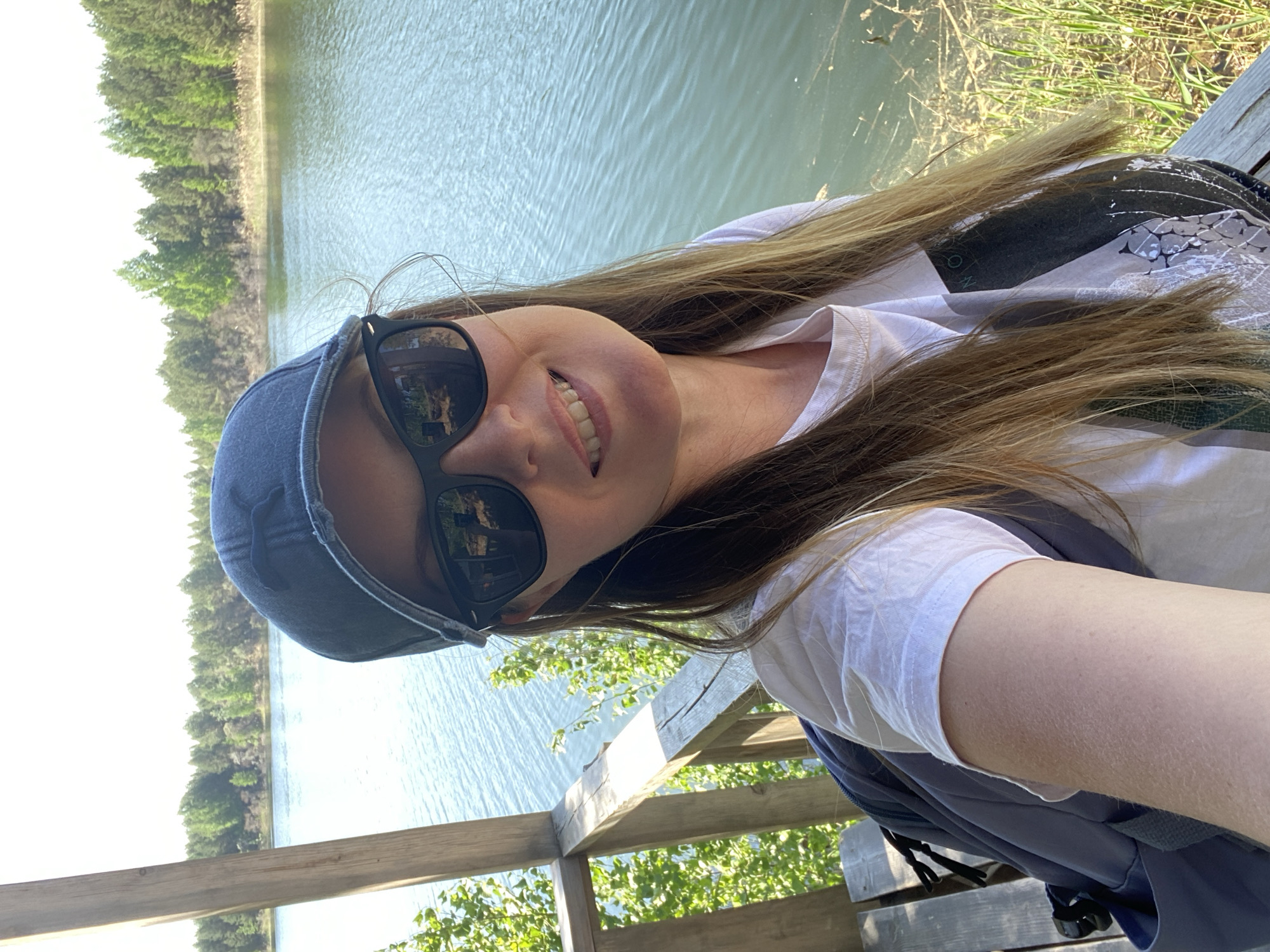 A woman is taking a selfie outdoors. She is wearing a white t-shirt, a baseball cap, and sunglasses. The background shows a body of water with trees lining the opposite shore. The selfie is taken on a wooden structure, possibly a bridge or observation deck. The location appears to be Novyi, Russia, based on the prompt.