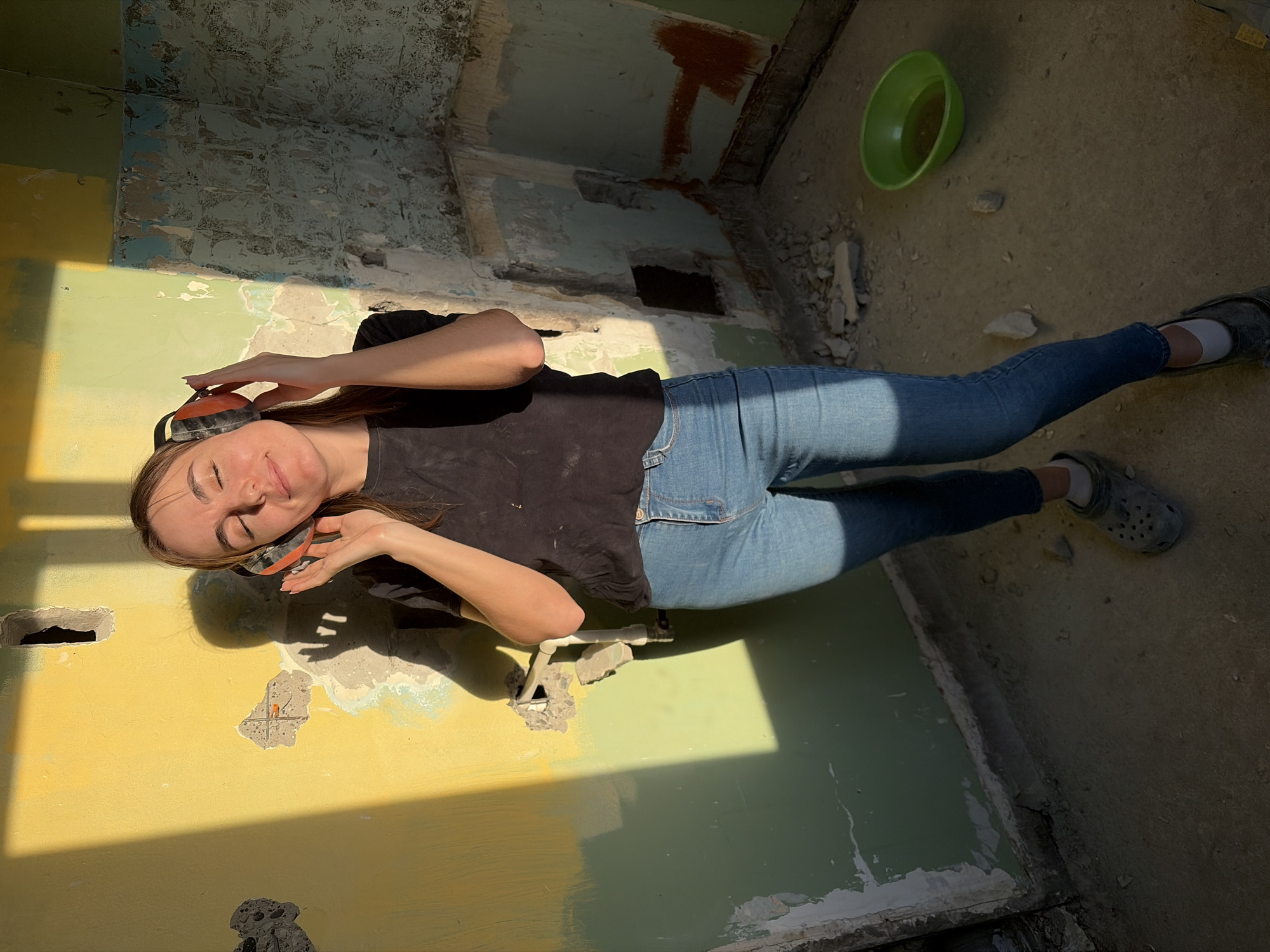 A woman with brown hair is lying on her side in the corner of a room undergoing renovation. She is wearing blue jeans, a black t-shirt, safety goggles on her forehead, and headphones around her ears. Her eyes are closed and she appears to be smiling.
The room has damaged walls with exposed concrete and layers of peeling paint in various colors, including yellow, green, and blue. There are holes in the wall, and debris is scattered on the floor. A green bowl sits on the floor in the upper right corner. The floor is dusty and cracked.
The location is unspecified, but it is not in Izhevsk, Russia.