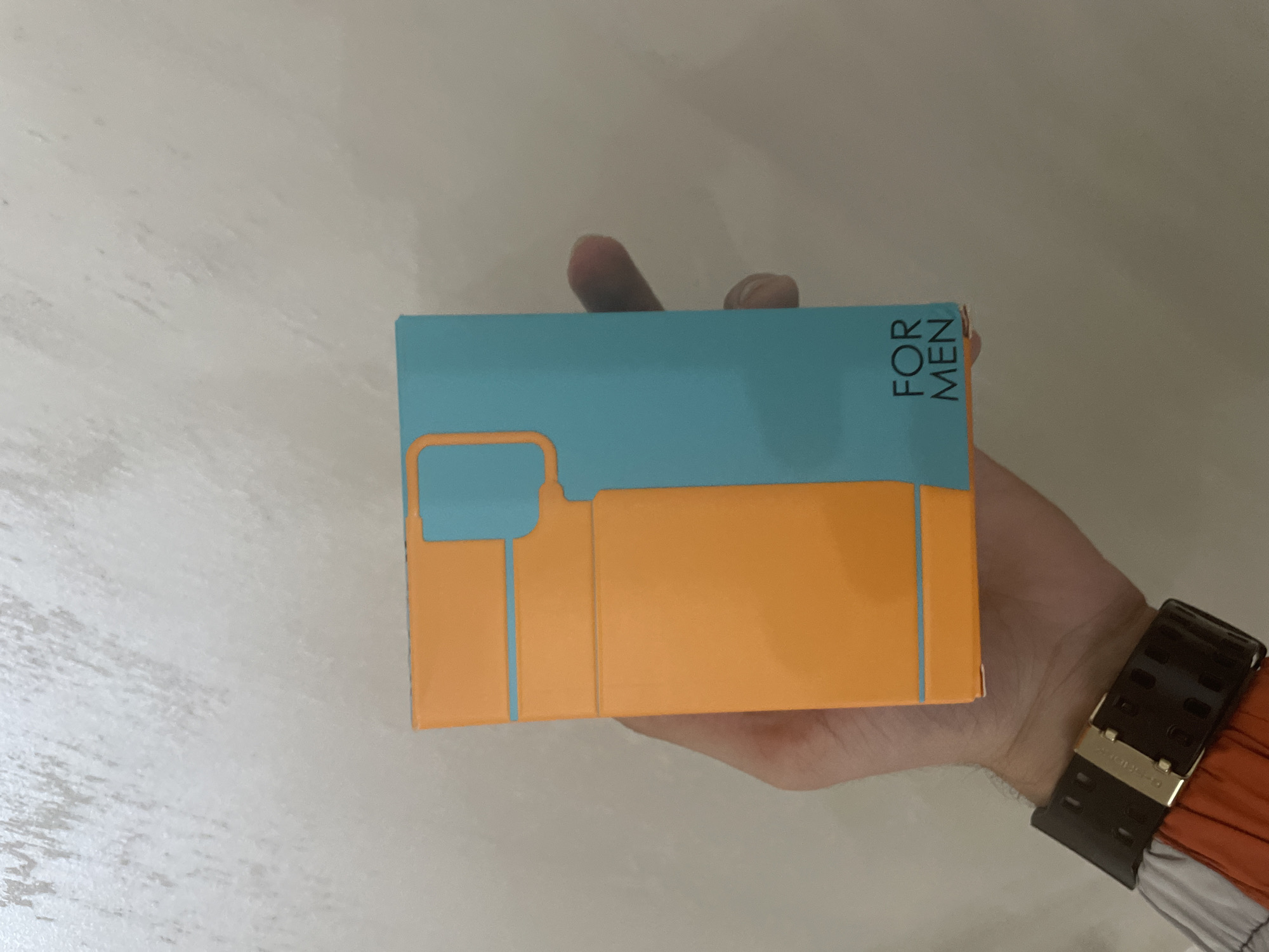 A close-up photograph captures a person's left hand holding a rectangular product box vertically against a plain, light-colored background. The box features a geometric design, divided between a bright orange section on the left and a teal section on the right, with orange lines forming an abstract representation of a bottle and cap. Black capital letters on the lower right of the teal section read "FOR MEN." A dark-colored wristwatch with a gold-toned buckle is visible on the person's wrist, and a portion of a sleeve with orange and white/grey stripes is visible on the lower left. The background is an unadorned light wall with a subtle, slightly textured finish. The image contains no visual elements or identifiers that indicate its location in the city of Ihtiman, Bulgaria, or any other specific geographical context.