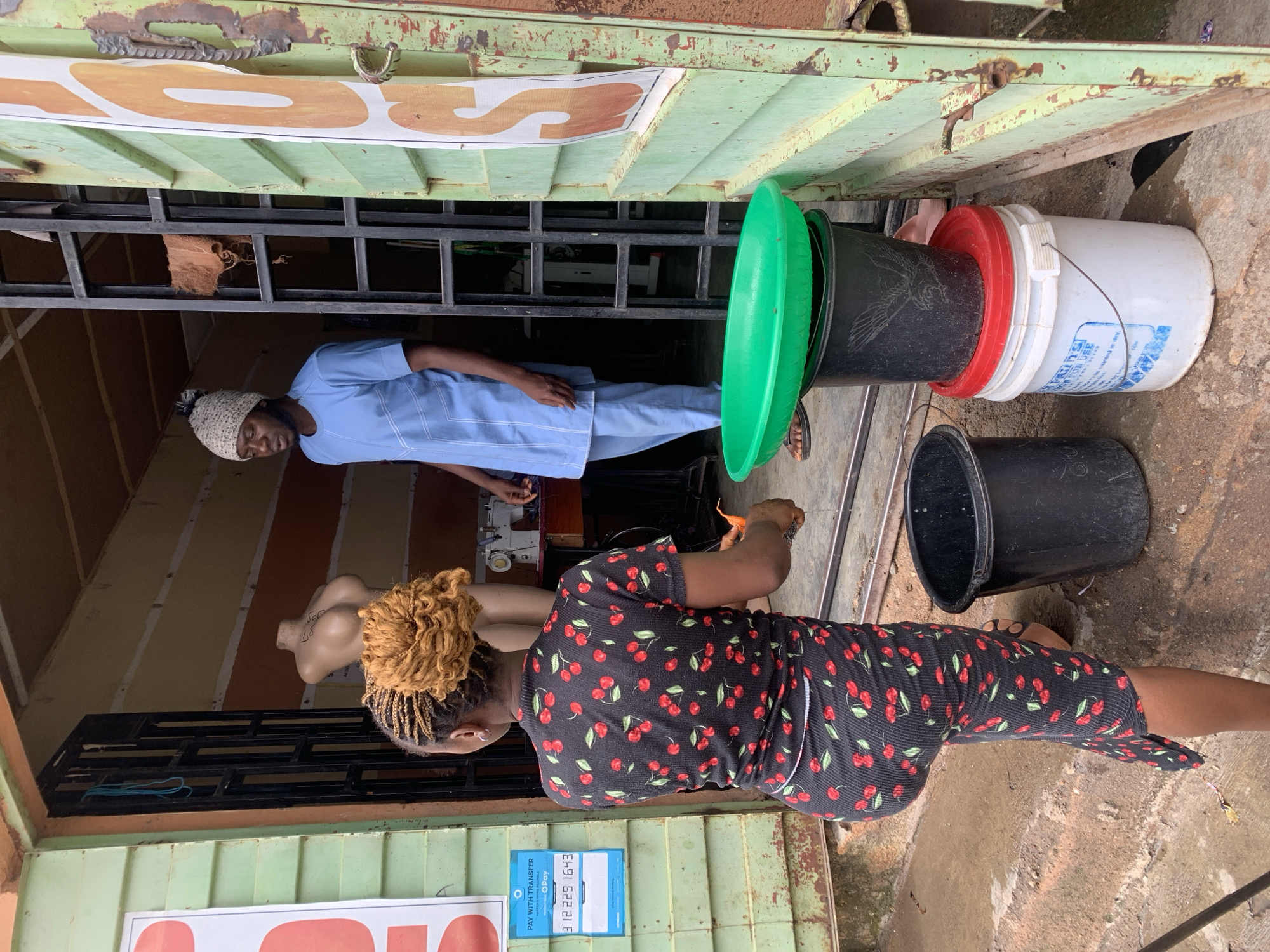 In Jos, Nigeria, a young Black woman with braided hair styled in a bun, wearing a black dress with a cherry print, stands facing away from the camera. She appears to be reaching down with her right hand. Beside her, a Black man in a light blue traditional outfit and a knitted cap with a pompom is visible inside a shop entrance. In the foreground, a stack of containers is visible: a white bucket with a red lid, a black bucket with a bird design, and a large green oval basin on top. To the left of the woman, a light green corrugated metal wall of the shop is visible, adorned with a white sign displaying the OPay logo and a phone number. Further inside the shop, a sewing machine can be seen on a table. The ground appears to be concrete and slightly wet.