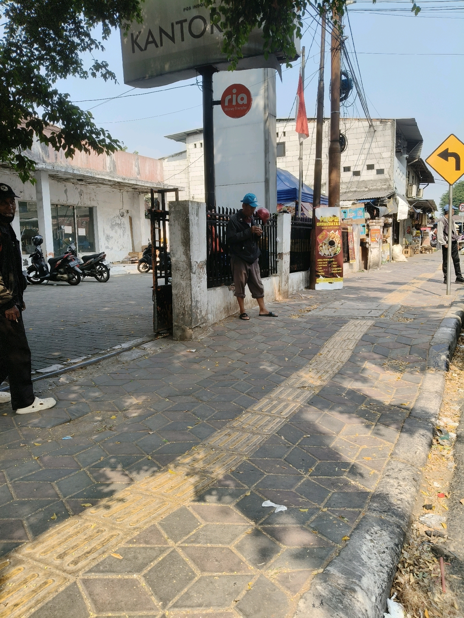 The image shows a street scene in South Tangerang, Indonesia. On the left, a person in a black uniform and black pants stands with their left foot forward. A row of motorcycles is parked behind them. In the center, a man wearing a blue cap, dark jacket, and grey pants stands looking at his phone, holding a red object. Behind him, a sign for "ria Money Transfer" is visible. Further to the right, a street vendor area is set up with stalls and goods. A yellow road sign indicating a sharp left turn is visible on the right side of the street. The sidewalk is paved with textured tiles, some of which are yellow to guide the visually impaired. Fallen leaves are scattered on the sidewalk and street, and there is some trash along the curb. The sky is clear and blue.
