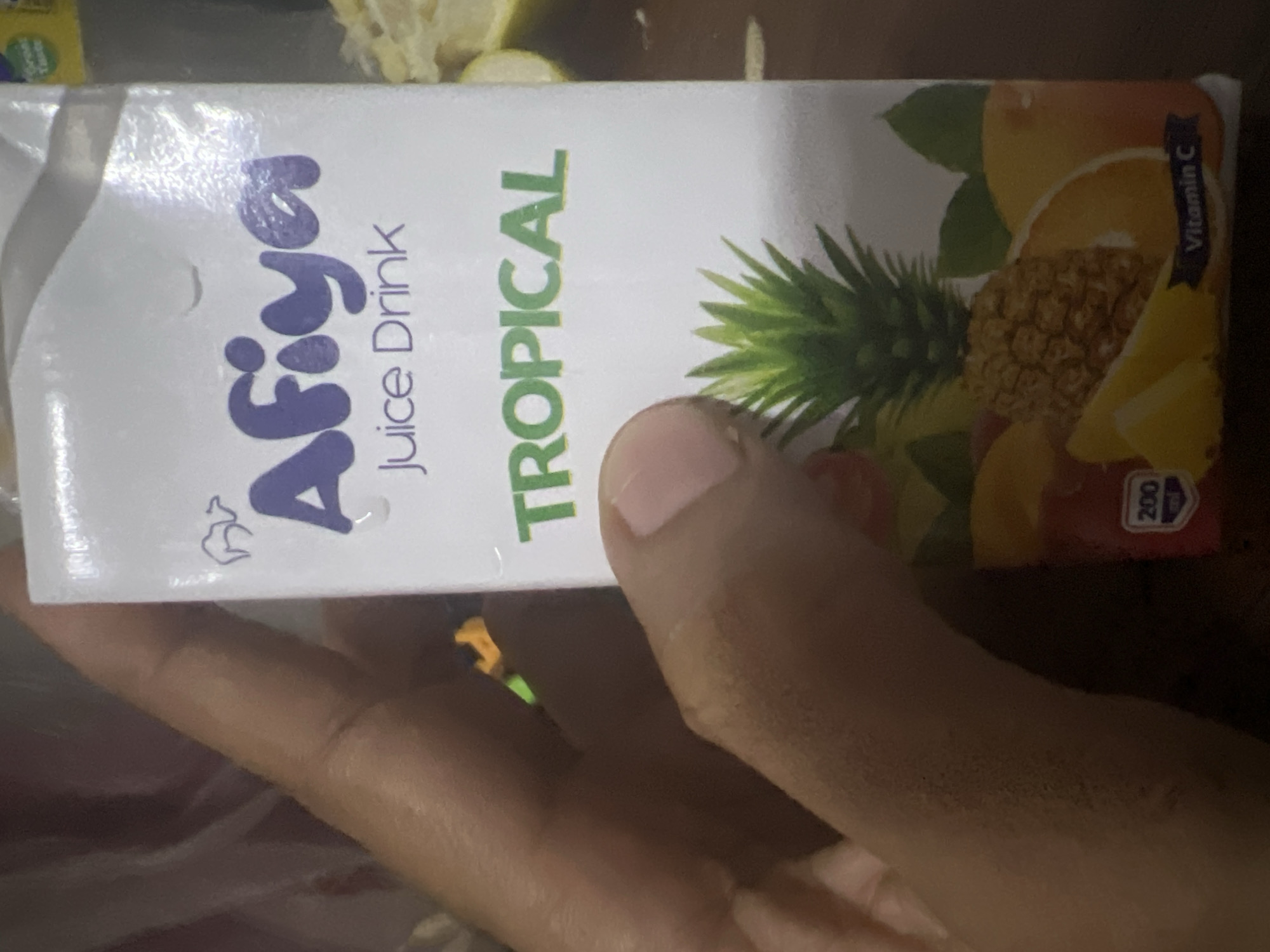 A hand holds a box of Afiya Tropical Juice Drink, which has the words "Juice Drink" on the box. The flavor is indicated to be tropical on the front and lists vitamin C on the right side of the box. The hand partially obscures the view of the juice box. The photo's setting is not clear from the given image, and there is no way to confirm its location within Dar es Salaam, Tanzania, or any other city.