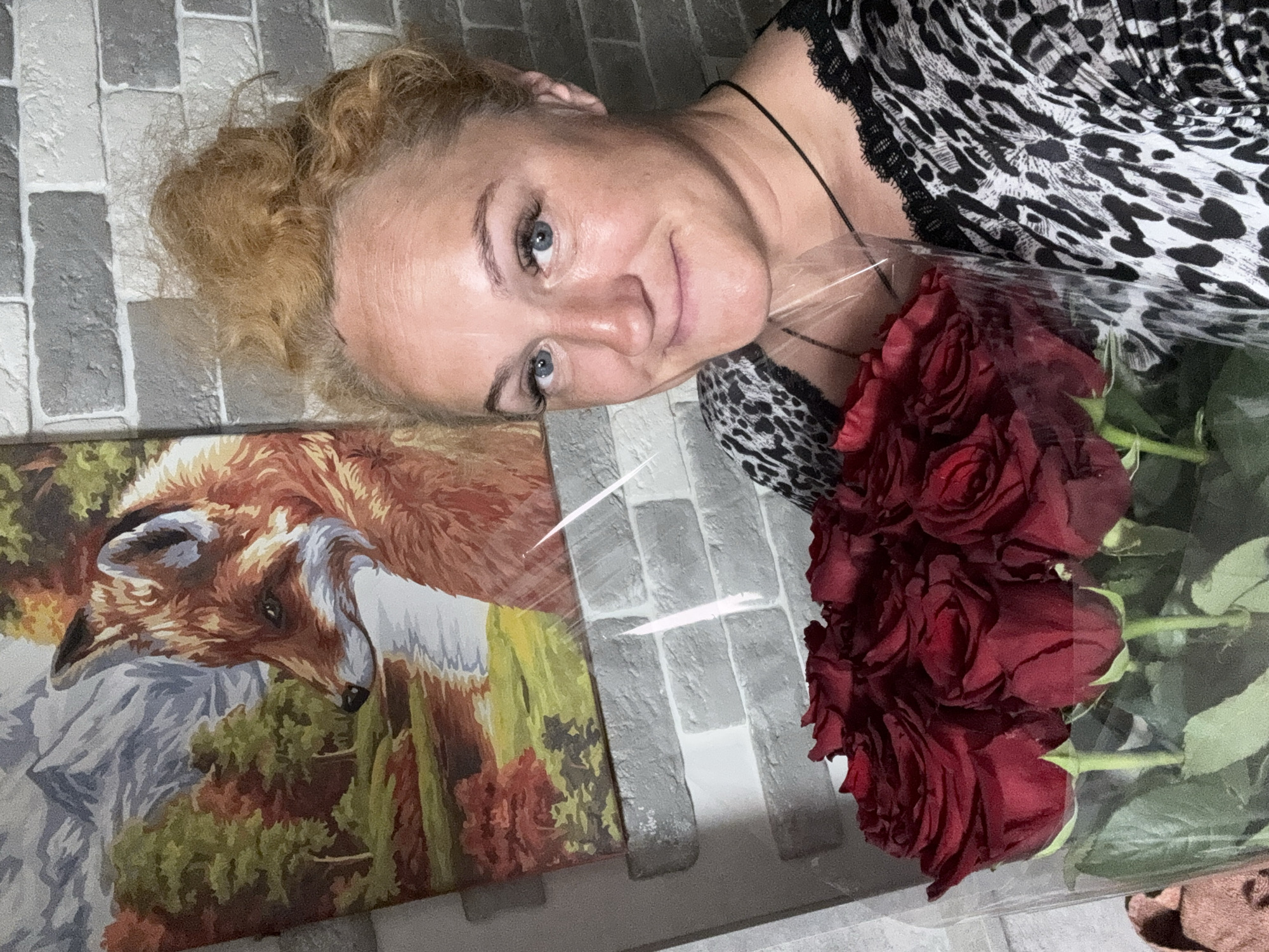 In a domestic indoor setting, a woman with blonde, messy hair tied up in a bun, blue eyes, and light skin, is holding a bouquet of red roses. She is wearing a black and white leopard-print top with lace trim. Behind her, a painting of a fox in a forest with mountains in the background hangs on a brick-patterned wall. The scene appears to be in a residential dwelling in the city of Chaikovsky, Russia.