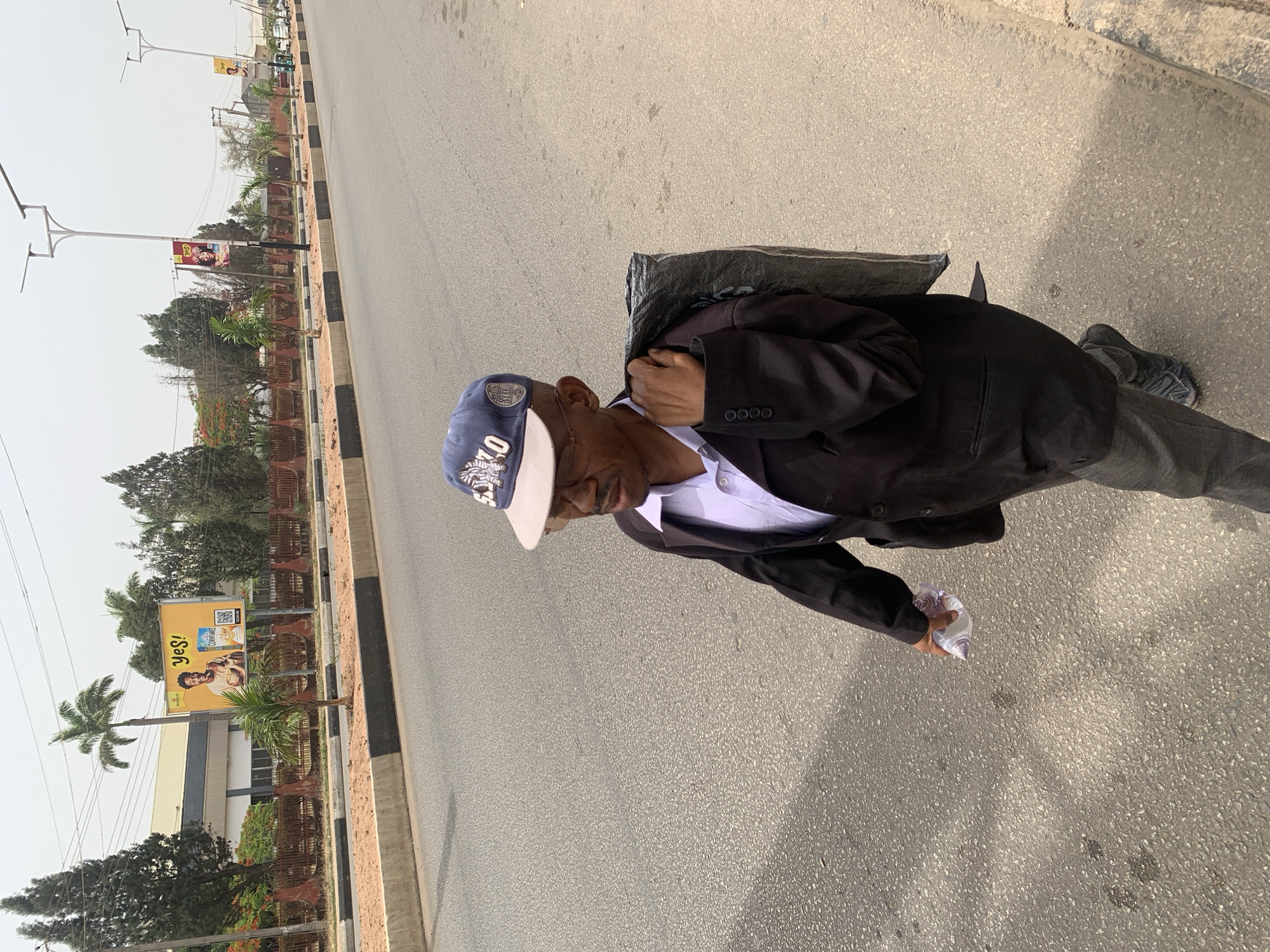 An elderly man is captured walking along the side of a paved road under bright daylight. He is dressed in a dark suit jacket over a light-colored collared shirt, grey trousers, and dark sneakers. A blue baseball cap with white lettering, appearing to spell "OILERS" and a number or initials like "07" or "LO", is worn on his head. He also appears to be wearing a light-colored necklace or pendant.
The man carries a dark bag, possibly a backpack or messenger bag, slung over his shoulder, and holds a crumpled clear plastic bag in his right hand. He is walking on the shoulder of a road that features a central median strip, adorned with small trees, shrubs, and decorative barriers or planters.
In the background, beyond the median, another lane of traffic is visible, followed by a roadside area with more trees and buildings. Tall streetlights with multiple arms line the road. Two billboards are visible: one prominently displays "YES!" and includes "FARNACO" at the bottom left, along with a QR code and an image of a person. The text "LUXON" or a similar word is also discernable on this billboard. The overall scene suggests an urban or semi-urban environment in Jos, Nigeria, characterized by clear skies and strong sunlight.