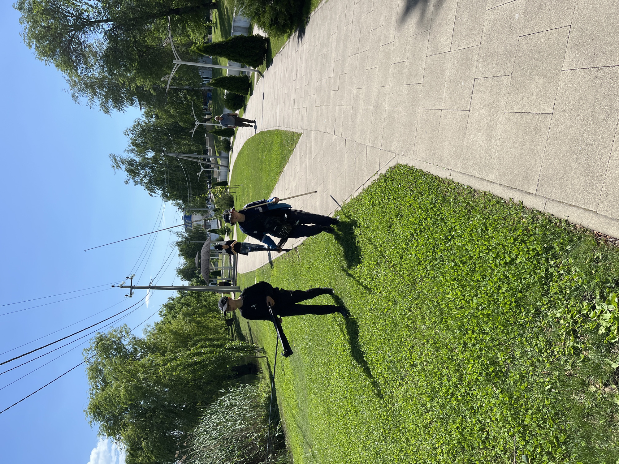 The image shows a park in Goryachiy Klyuch, Russia. A paved pathway curves through the scene, bordered by green grass. Two individuals in dark clothing are visible, one holding a long pole. Power lines run overhead. Trees and other greenery surround the park. In the background, another individual is walking on the pathway.