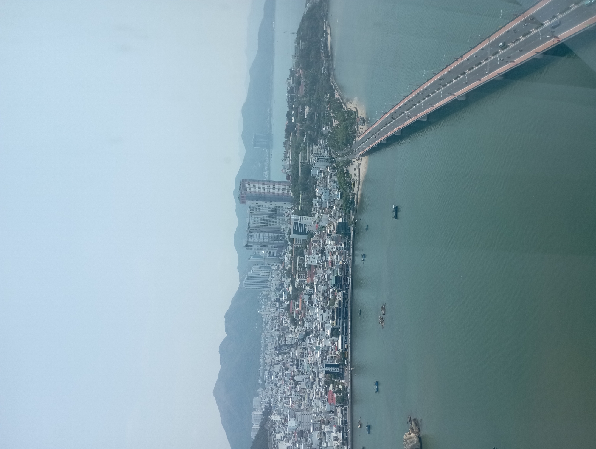 The image shows an aerial view of a city with a long bridge extending into the ocean. The city, presumed to be Nha Trang Ward in Vietnam, is nestled between mountains and the sea, featuring a dense urban landscape with numerous buildings, including several high-rises. The bridge, a significant structure with multiple lanes, connects the mainland to a narrow strip of land extending into the water. There are boats visible in the ocean near the city. The weather appears hazy, and visibility is limited. The image captures a broad, sweeping view of the urban environment and coastal landscape.