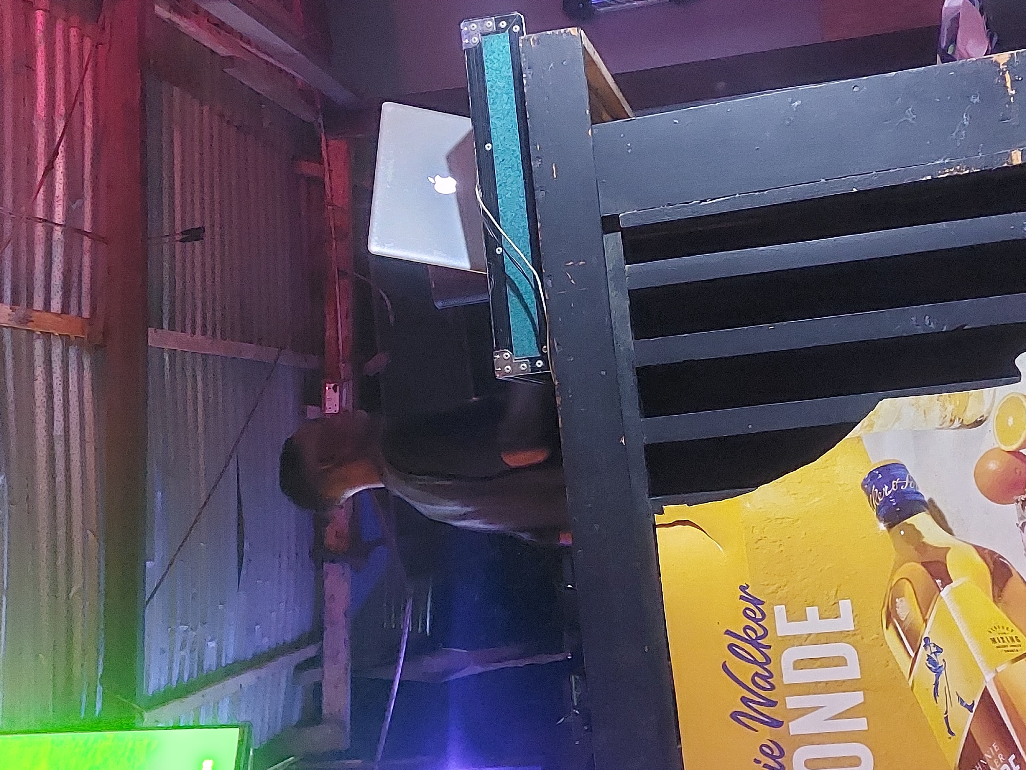 The image shows an interior space in Dar es-Salaam, Tanzania. A man is present, although his face is partially obscured. He is wearing a dark shirt. A laptop with an Apple logo is affixed to a vertical structure made of dark wood, which resembles a stand or shelf. The background includes corrugated metal walls, illuminated by colored lights (red, blue, and green). A large advertisement for Johnnie Walker Blonde whiskey is also visible, partially covered by the wooden structure.