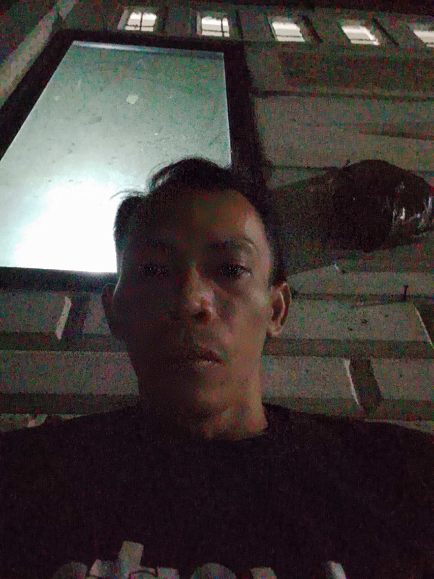The image depicts a person with dark hair and a dark shirt in an interior setting. The person appears to be facing the camera. Behind the person, there is a bright, reflective surface, possibly a window or a screen. Above the reflective surface are several window openings. The background shows a textured surface with horizontal lines, possibly a building facade. Based on the instructions, the location is Bukit Ketok, Indonesia, but there is no concrete detail in the image to confirm this.