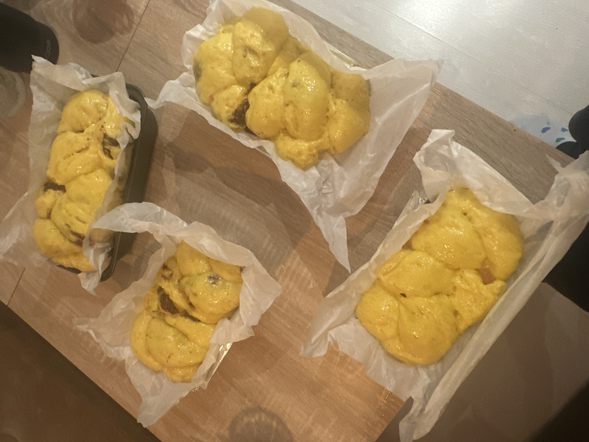 On a wooden surface are four yellow buns arranged in white parchment paper. One bun is in a dark metal pan, the other three are simply set on the parchment. No humans are visible. Location is unknown.