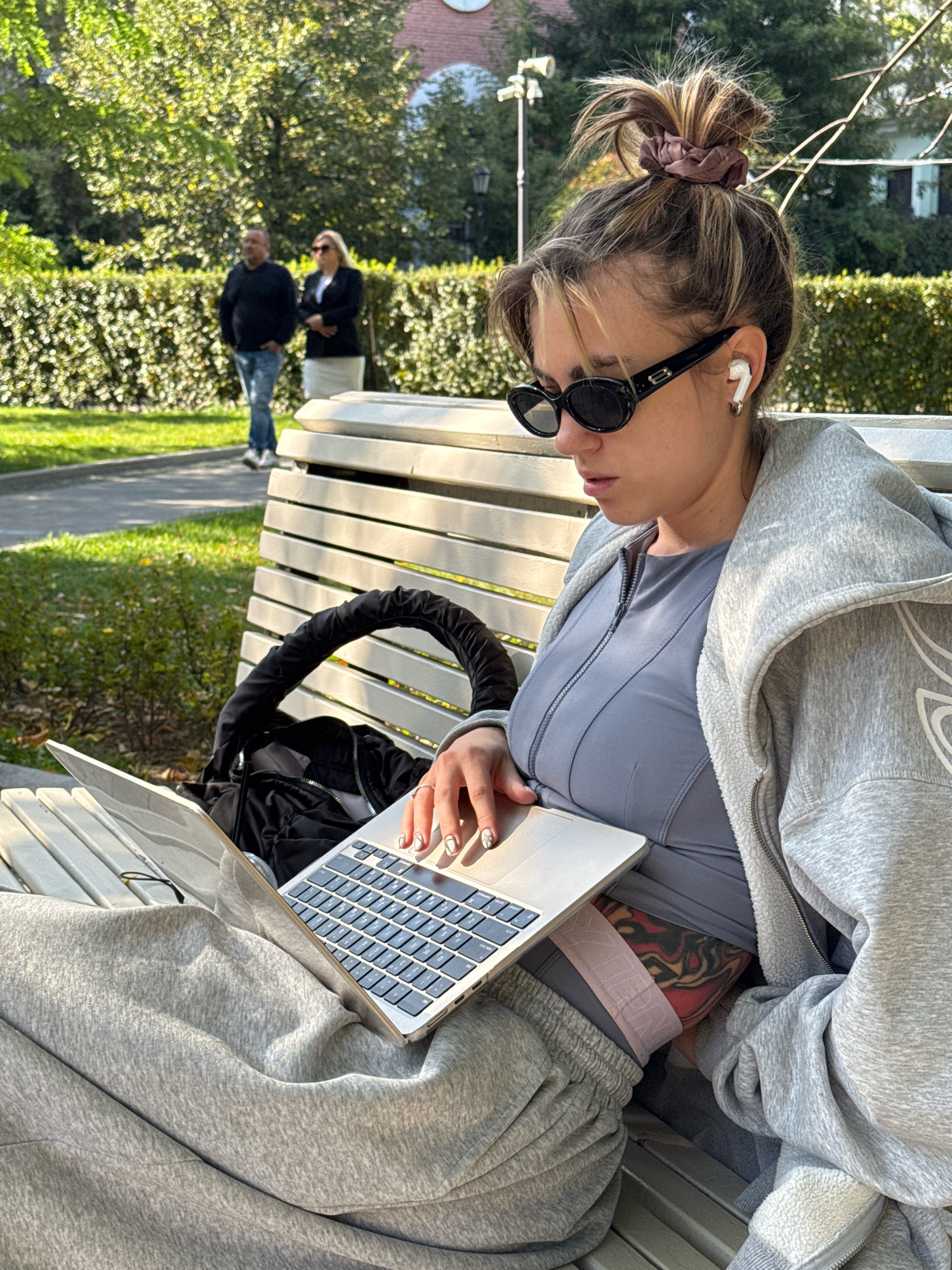 In Moscow, Russia, a young woman with blonde hair pulled up in a messy bun, wearing sunglasses and earbuds, sits on a park bench with a laptop. She is dressed in a grey hoodie and grey athletic pants, with a grey fitted top underneath. Her fingernails are painted with a white and grey marbled design. A tattoo is visible on her hip. A black backpack is on the bench behind her. Two people, a man and a woman, walk in the background. The setting appears to be a park with trees and a manicured hedge. Sunlight illuminates the scene.