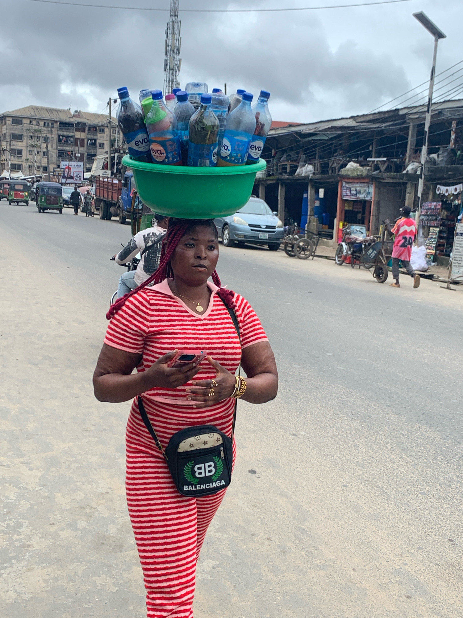 In Umuode, Nigeria, a woman walks along a paved road, carrying a large green bowl filled with bottled water on her head. She wears a red and white striped dress and a black crossbody bag with a Balenciaga logo. Her hands are clasped in front of her, and she holds a mobile phone. In the background, several vehicles, including a truck, tricycles, and a car, are present on the road. Buildings line the street, some appearing commercial with awnings and displays of goods. The sky is overcast with grey clouds.