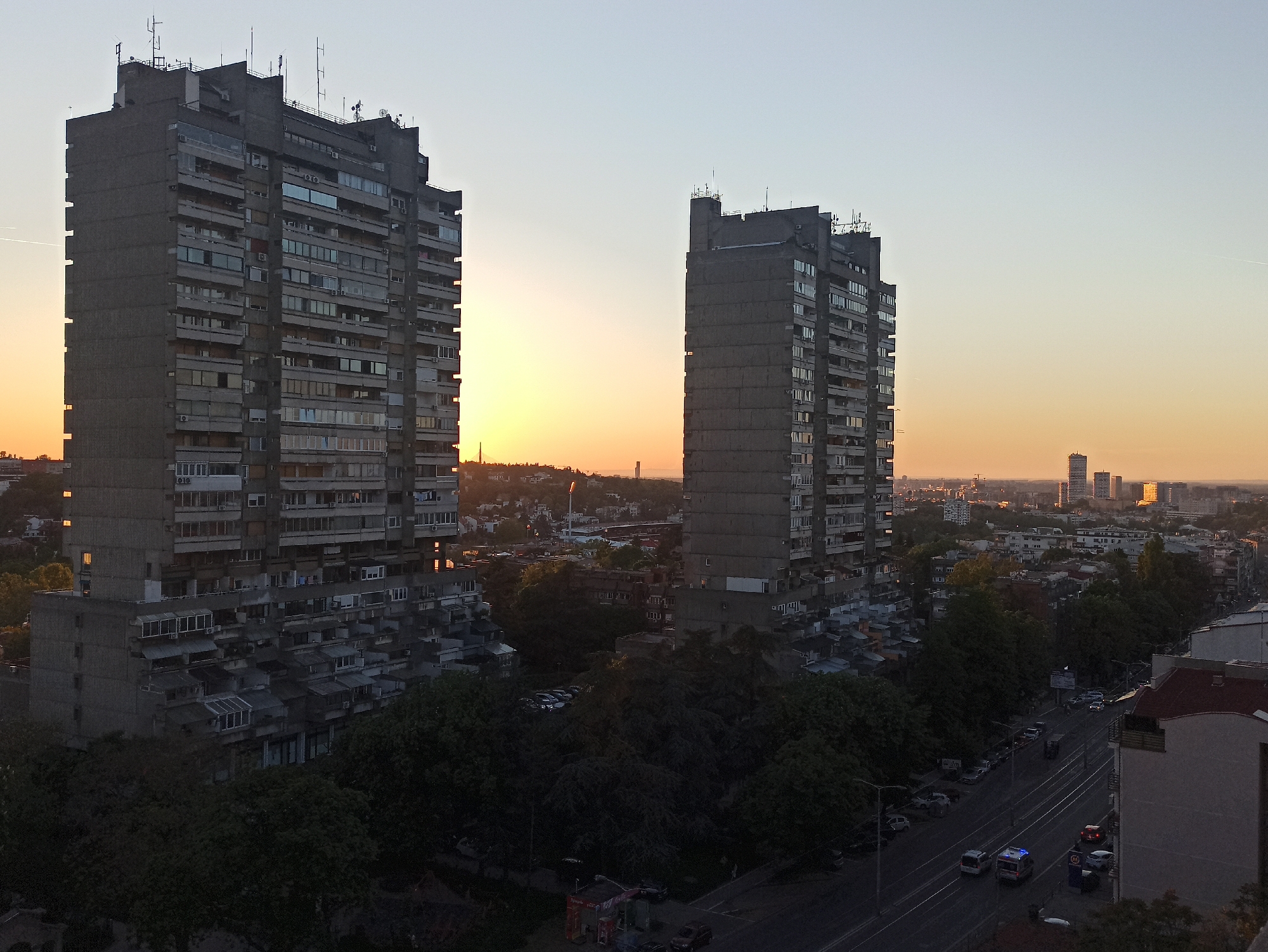 The image shows two tall, grey concrete buildings, likely residential, in Vozdovac Urban Municipality, Serbia, during a sunset. The sun is positioned between the two buildings on the left. There is a city landscape with trees and other buildings extending into the background. A street with a few parked cars and some moving vehicles (one with blue lights) is visible in the foreground.