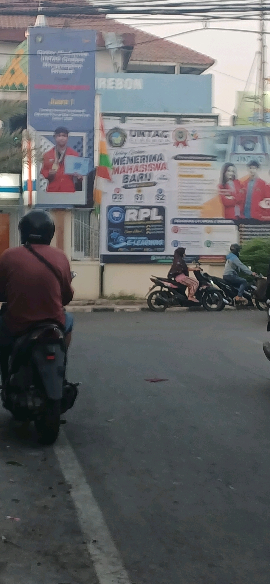 The image shows a street scene in Cirebon, Indonesia, with several billboards visible. One billboard announces that UNTAG Cirebon is accepting new students. Another billboard displays a young person holding a medal, possibly indicating an achievement in a competition. There are also people riding motorcycles on the street. The environment appears to be an urban area with buildings and overhead wires.