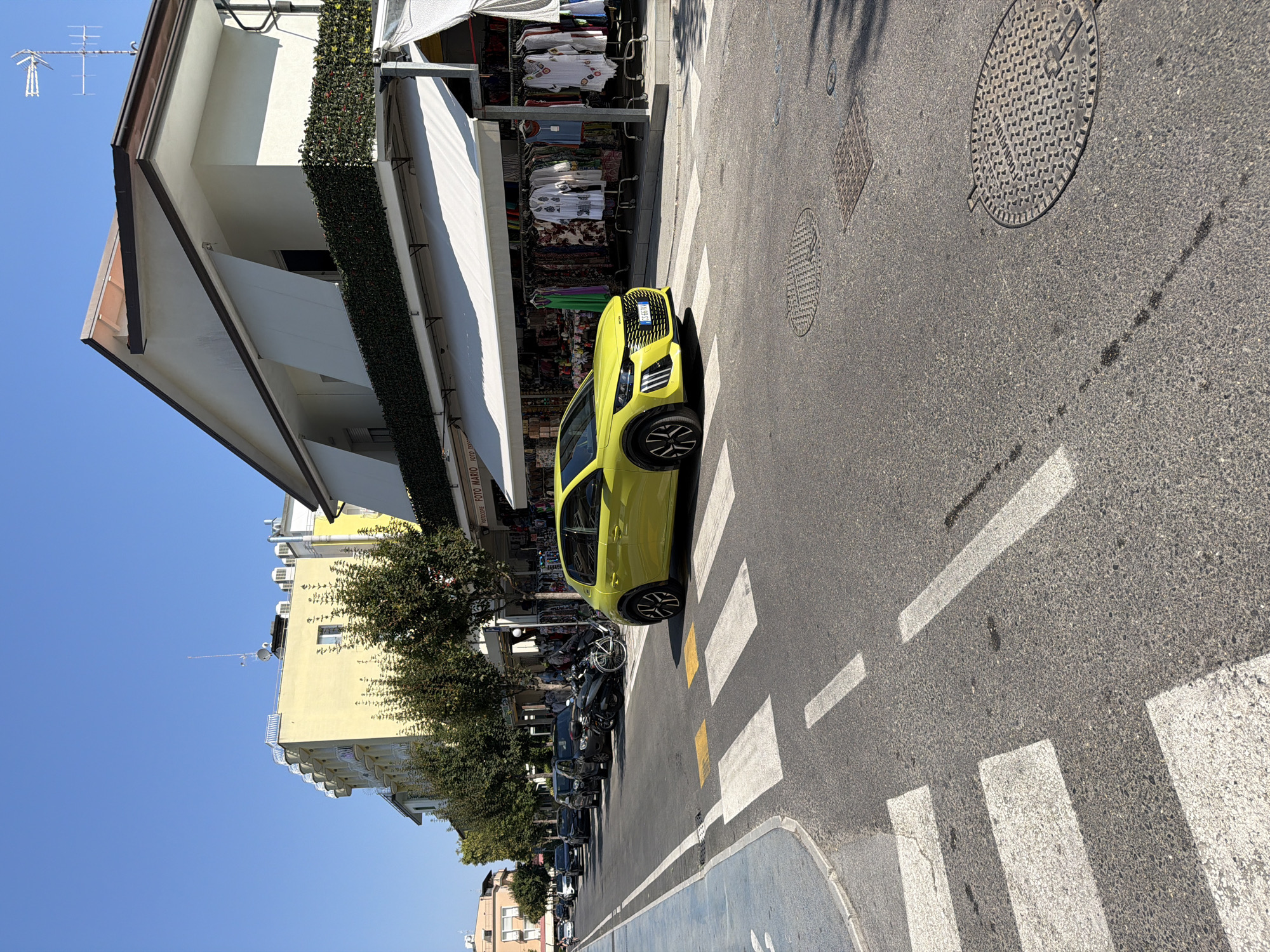 The image depicts a street scene in Gatteo, Italy, on a clear, sunny day. The foreground features a pedestrian crossing with white stripes on asphalt. A bright yellow car is positioned on the crossing, facing right. Beyond the crossing, the street is lined with parked cars and motorcycles. Shops with awnings are visible on the right side of the street, displaying clothing racks. In the background, buildings with multiple stories are seen, characterized by light-colored facades and balconies. Trees are planted along the sidewalk. The sky is a vibrant blue.