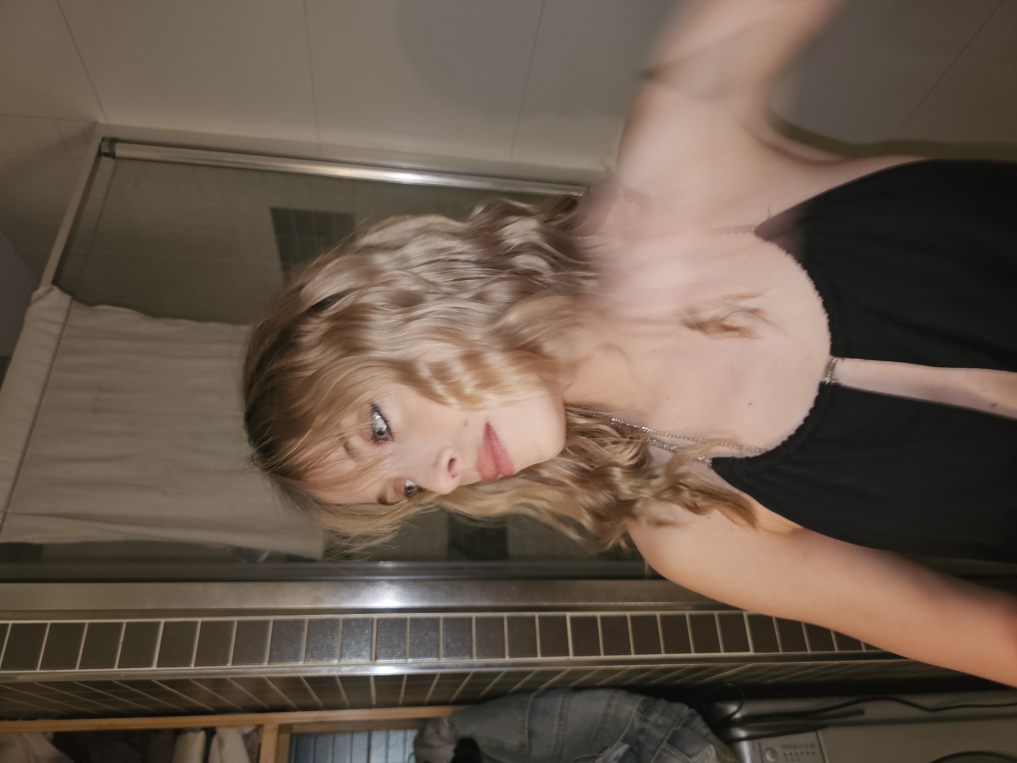 A young woman with blonde, wavy hair is shown in the image, wearing a black top with silver straps. She is facing slightly to the left, with her head tilted. Her expression is pensive, with her eyes looking downwards. A silver necklace is visible around her neck. The background includes a bathroom or laundry area, with a shower enclosure, tile details, and a glimpse of laundry.