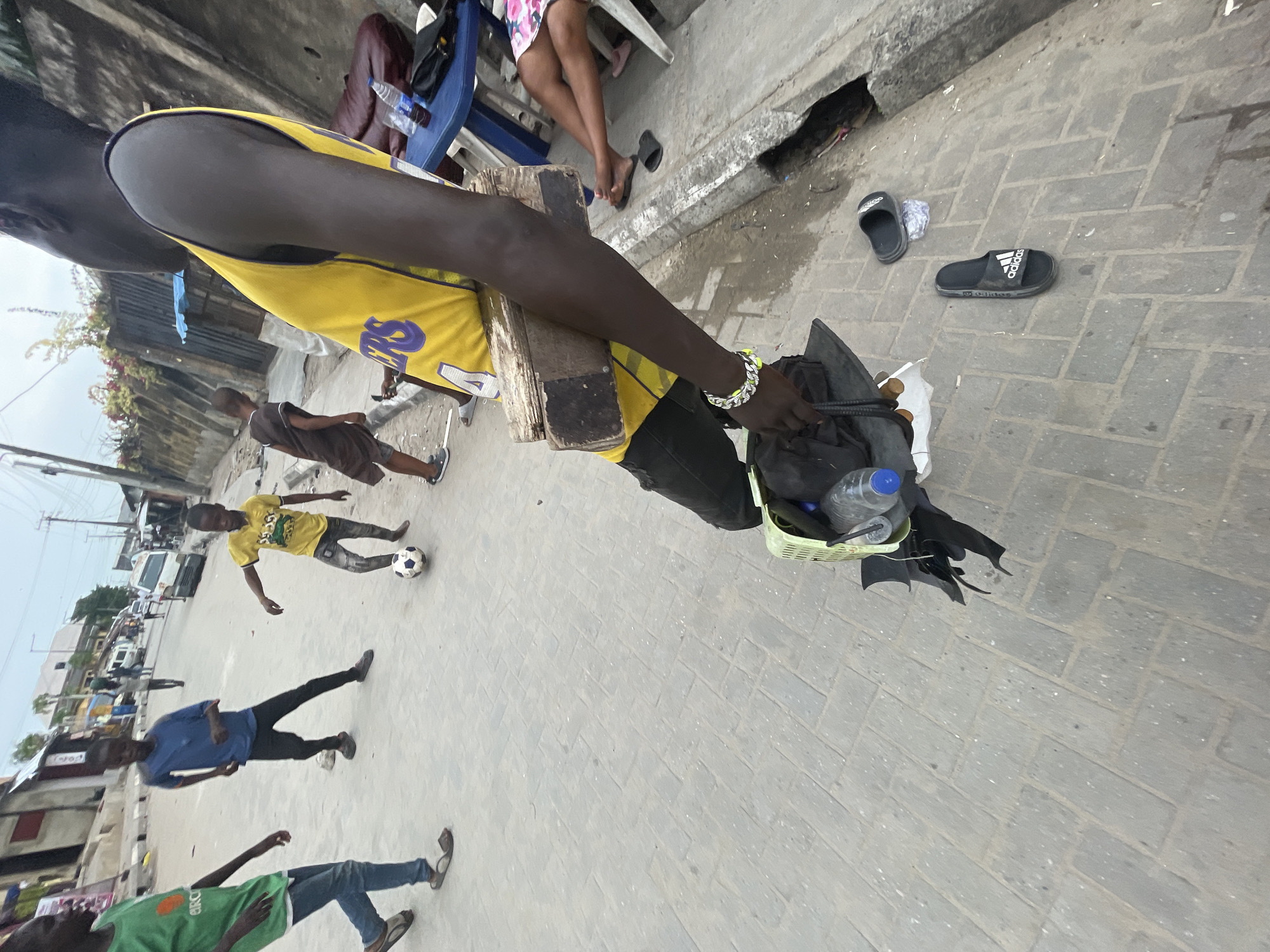 The image, taken in Ado, Nigeria, is rotated 90 degrees clockwise and depicts a lively outdoor street scene. In the prominent foreground, a dark-skinned individual wears a yellow and purple basketball jersey, appearing to be a Lakers jersey with the number "24" and "LAKERS" written vertically. They are holding a wooden stool or platform with their left hand, while their right hand reaches into a small green plastic basket containing clear water bottles and other items, suggesting they might be a vendor. A silver and yellow bracelet is visible on their left wrist.
The ground is paved with grey interlocking bricks. To the right of the main subject, a pair of black Adidas slides are visible, with the "adidas" logo clearly discernable on one. Further back and to the right, two pairs of adult legs are seen, one wearing sandals and a floral skirt, the other in dark clothing, seated around what appears to be a small table with a blue plastic chair.
In the mid-ground, several children and young men are present. One child in a yellow t-shirt and blue jeans is standing near a black and white soccer ball, seemingly playing. Another child in a brown t-shirt and shorts is further back. Two other young men are also visible: one in a blue t-shirt and black pants, and another in a green t-shirt with partial text "CIRCL" visible, wearing blue jeans.
The background shows glimpses of buildings, power lines, and possibly parked vehicles, indicative of an urban or semi-urban street. The lighting suggests it is daytime, with bright but diffuse light, possibly on an overcast day or during late morning/afternoon. The overall scene is a candid snapshot of daily life, with people engaged in various activities on a street.
