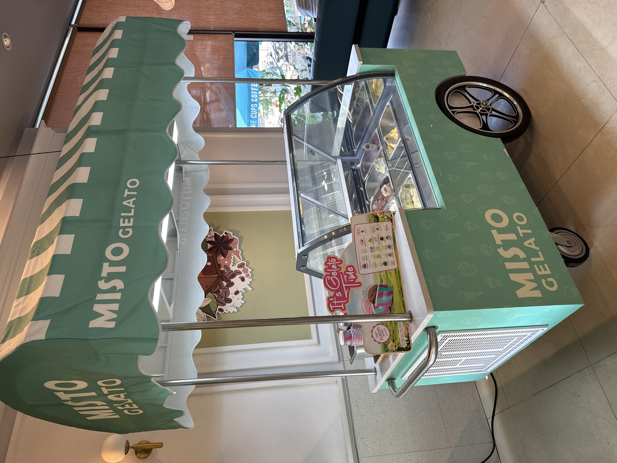 The image shows a "Misto Gelato" cart, likely indoors given the flooring and partial view of a window with the text "HE CUPS COFFE," indicating a cafe or similar establishment. The cart is light green with white lettering and is adorned with a striped awning bearing the brand name. The cart contains a glass-enclosed display of various gelato flavors. There is a menu board with prices and images of gelato. A decorative graphic of coffee beans and chocolate is attached to the side. The cart has wheels for mobility. The cart appears to be inside near a wall where a light fixture is installed. A portion of a blue couch is visible. The location appears to be in Đà Nẵng, Vietnam, due to the Vietnamese text "OTAJOTKIM."