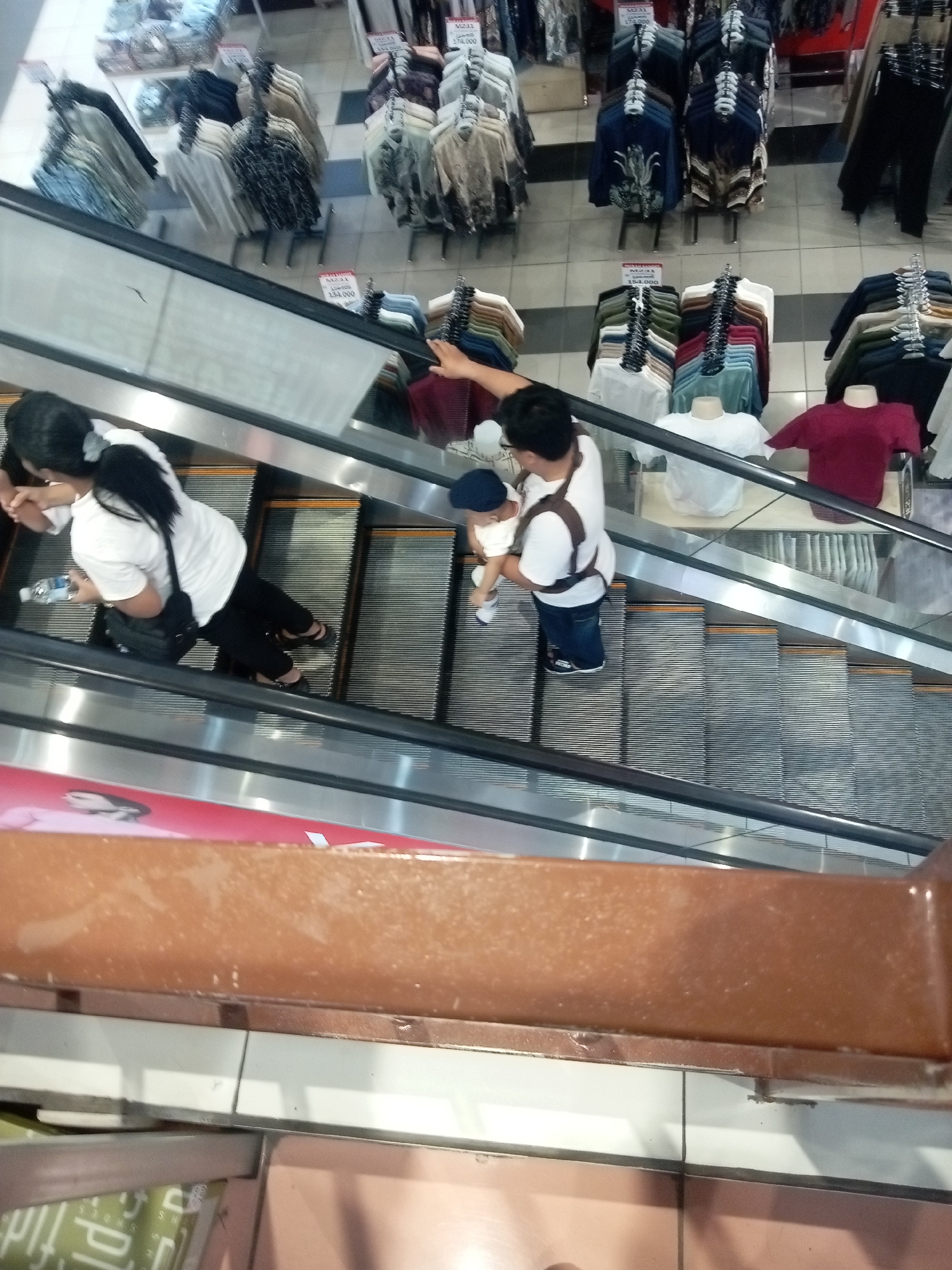 An overhead, slightly angled medium shot captures a view of an escalator within a department store, likely in Batam, Indonesia, given the context. The escalator's ribbed steps are visible, some with yellow and orange accents. In the foreground, a brown railing runs horizontally, showing dust and minor imperfections.
On the escalator, two primary subjects are present. On the left, a woman with dark hair, wearing a white t-shirt and black pants, is holding a black purse on her left hip. She has a water bottle in her right hand. Her posture suggests she is moving downwards on the escalator.
To her right, a man with dark hair, wearing a white t-shirt and a brown harness system, is carrying a baby. The baby is facing away from the camera and is wearing a light-colored outfit and a blue hat. The man is looking down at the baby, and both are on the escalator moving downwards.
In the background, rows of clothing are displayed on racks. These appear to be shirts and blouses in various colors, including blues, grays, whites, and reds. Some clothing items have price tags. The overall environment is well-lit, typical of a retail setting.