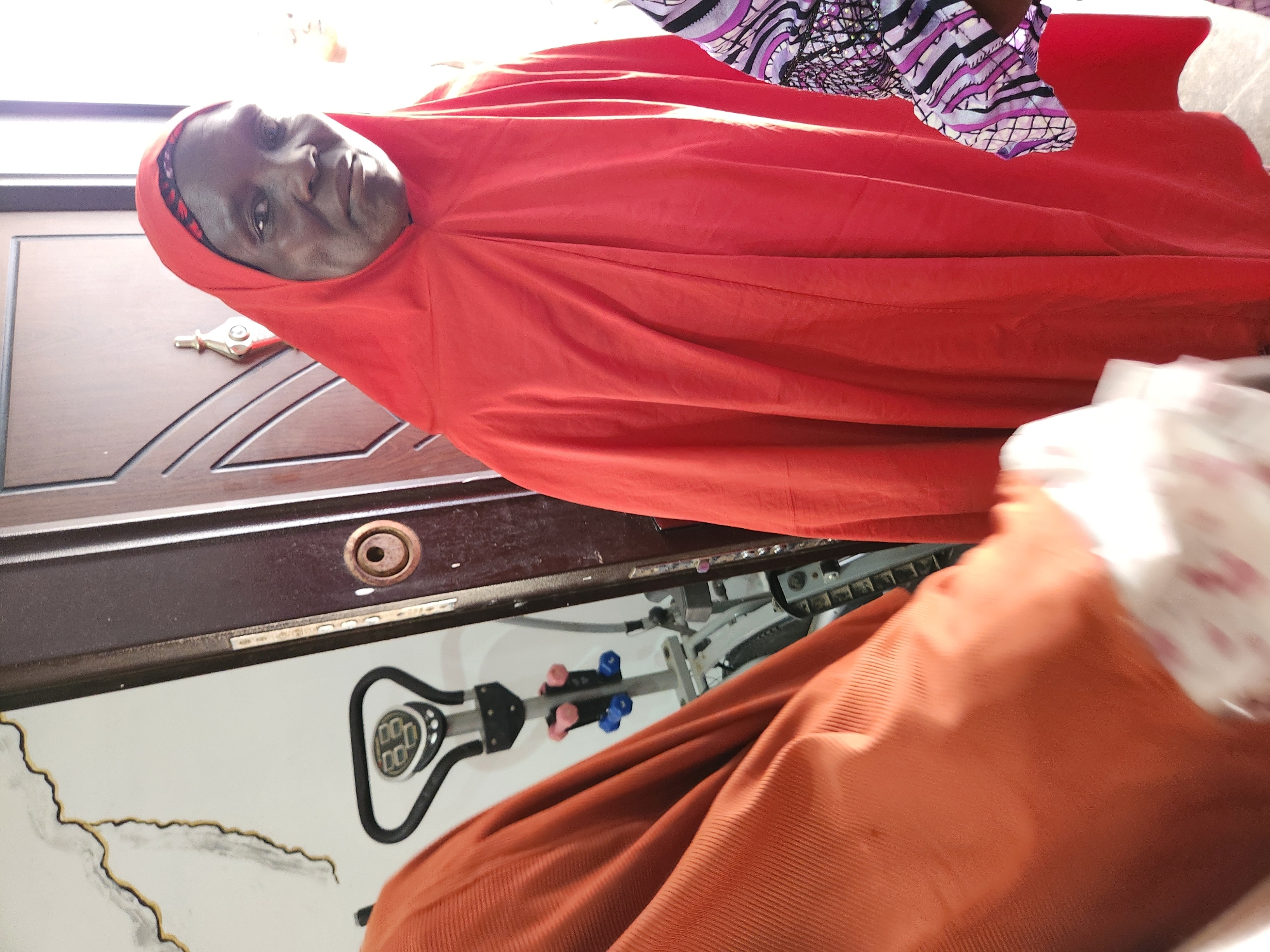 The image shows a medium shot of a woman wearing a red hijab and an orange dress. She is standing in front of a brown door. There is a piece of exercise equipment to her left, and a white wall with some black and white drawings behind it. Another person is partially visible on the right side of the image, wearing a purple and white patterned garment. The lighting in the image is bright, suggesting it was taken during the day. The context of Sabon Tasha, Nigeria is not discernible from the visual elements alone.